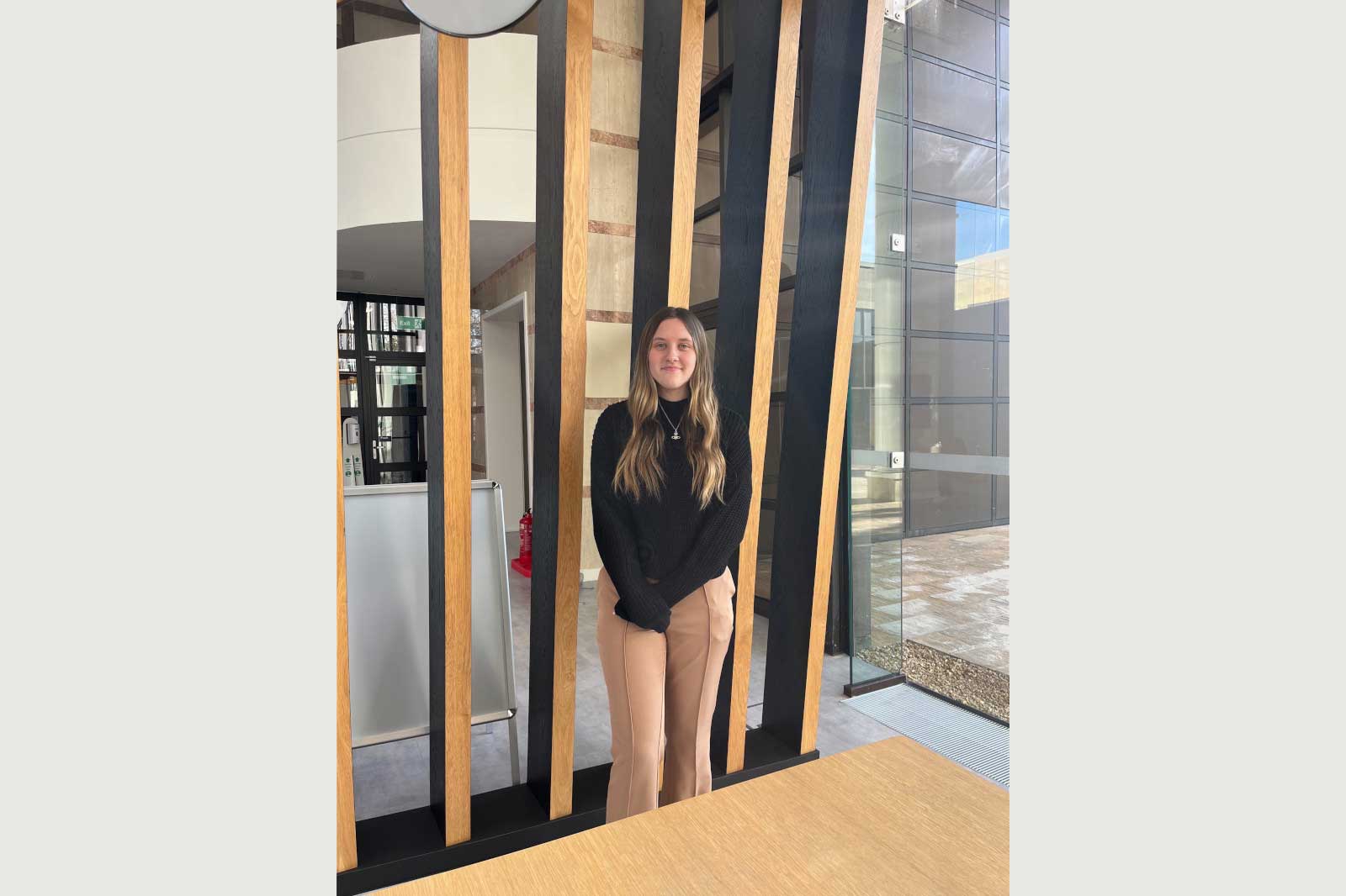 A person stands between wooden and black vertical slats in a modern interior with glass walls and a wooden table.