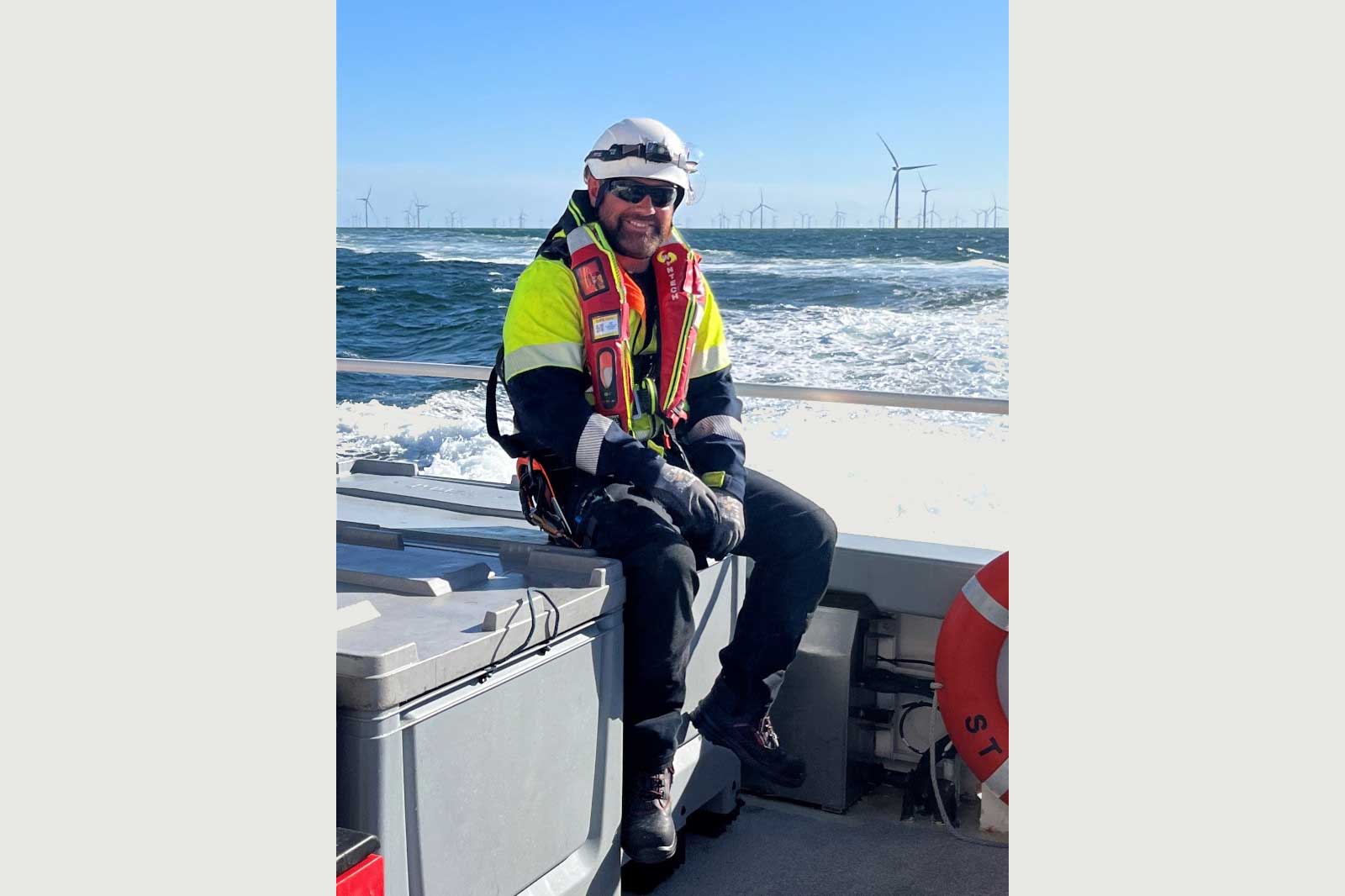 A person in safety gear sits on a boat, with wind turbines visible in the background and waves splashing behind.