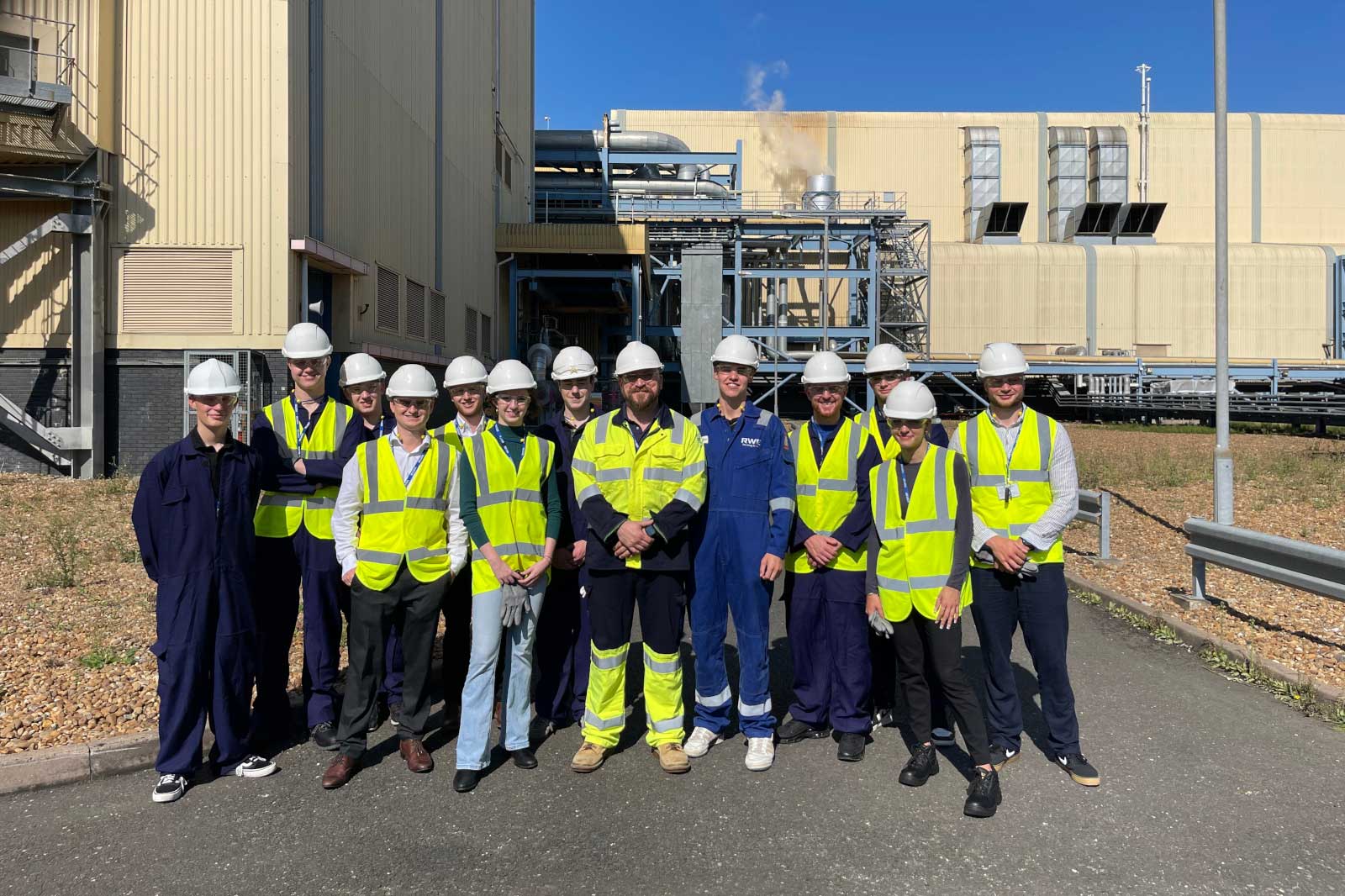 A group of workers in high-visibility vests and helmets standing outside an industrial facility under a clear blue sky.