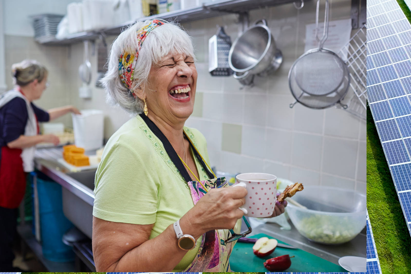 A woman holds a polka-dotted cup and stands in a kitchen preparing food, with another person working in the background.
