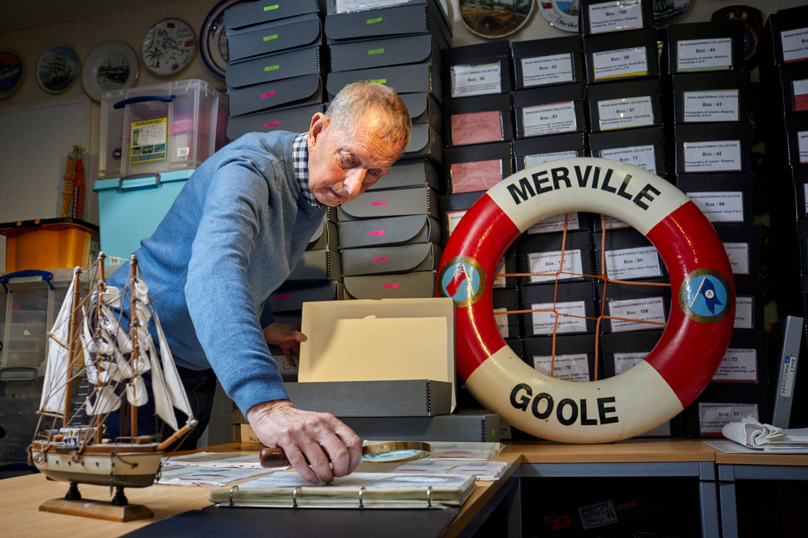 A person inspects a box on a desk surrounded by storage boxes, a ship model, and a decorative lifebuoy marked 'Merville Goole'.