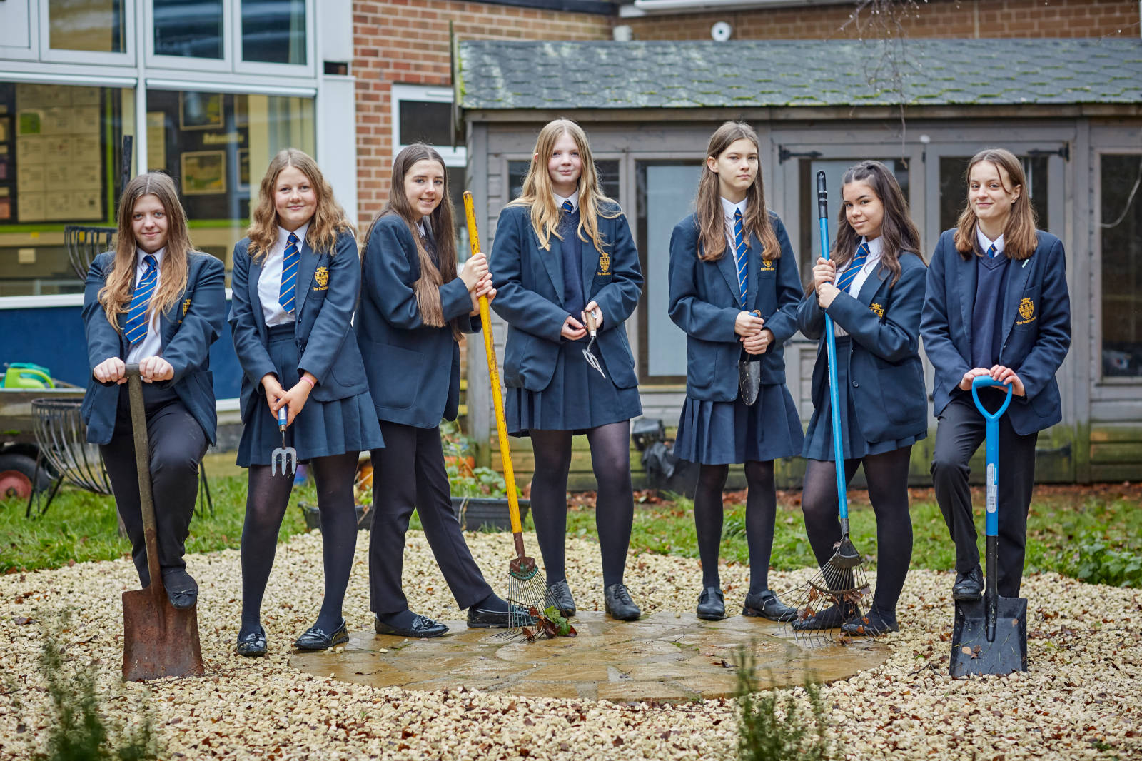 Seven girls in school uniforms stand with gardening tools on a stone pathway, surrounded by gravel and greenery.