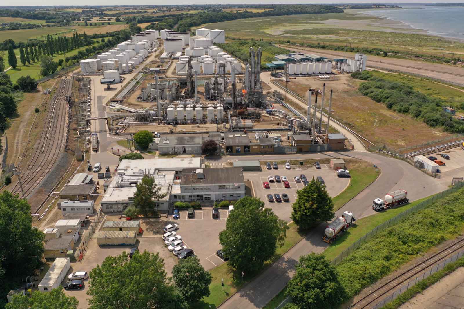 Aerial view of an industrial site with storage tanks, processing equipment, and a nearby railway, surrounded by greenery.