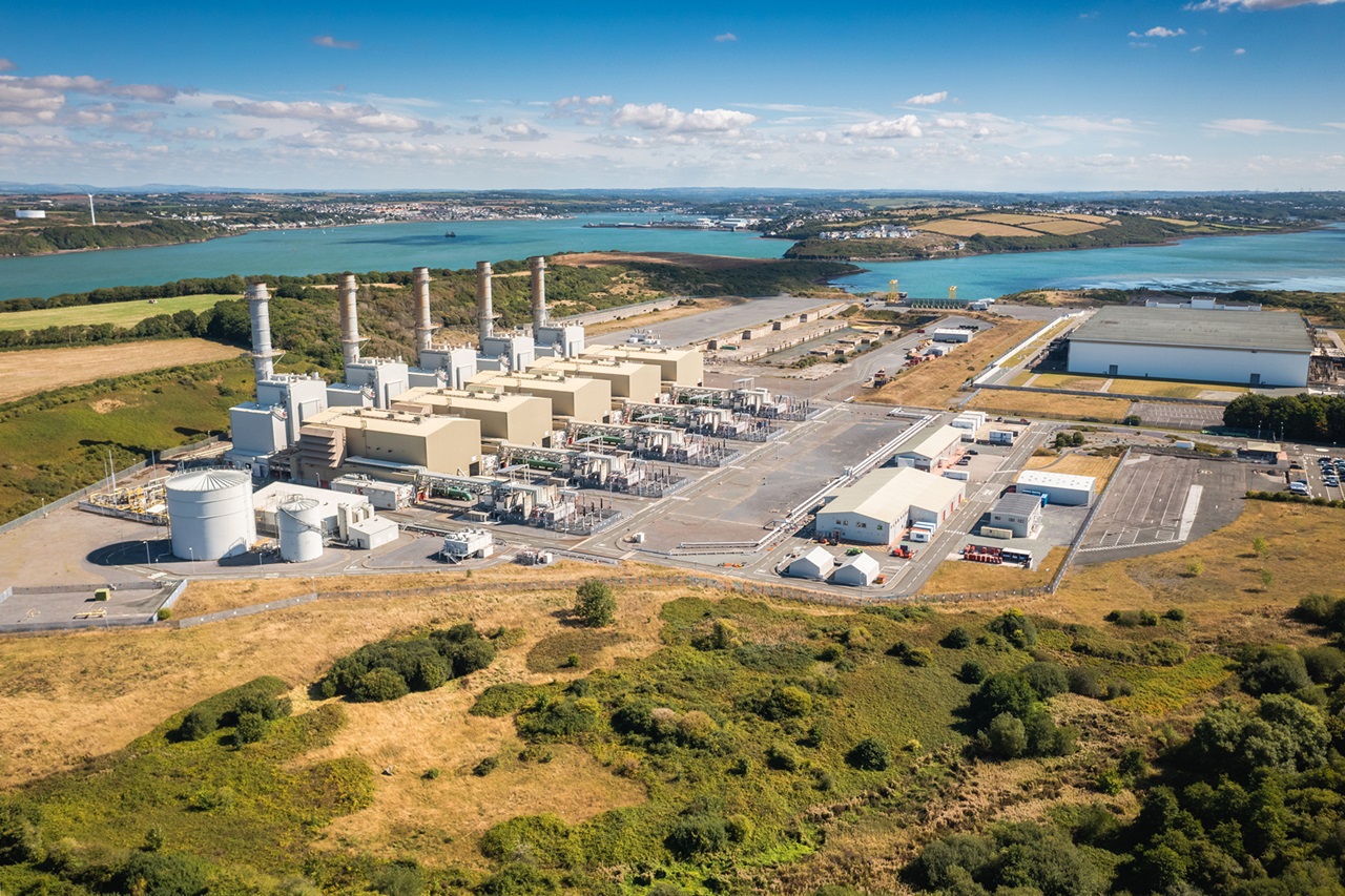 Aerial view of an industrial complex with gas turbines, storage tanks, and a scenic river in the background.