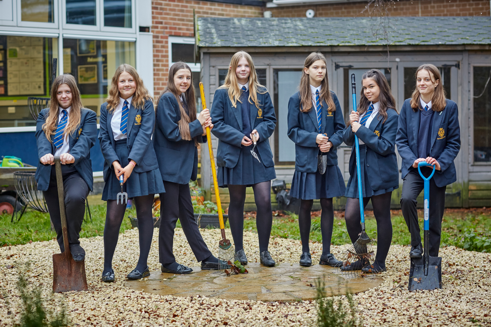 A group of schoolgirls in uniform stands outdoors with gardening tools on gravel. Behind them, there are windows of a building.