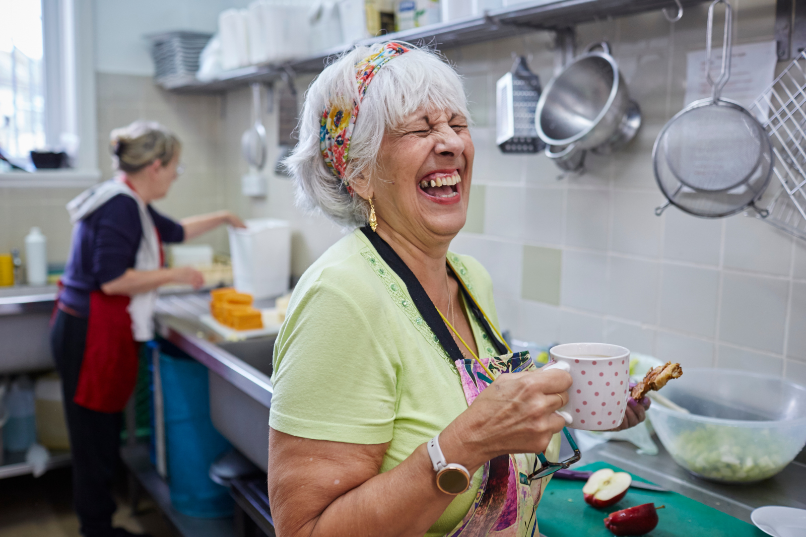 A woman holds a cup in a kitchen while another person works in the background, chopping vegetables and preparing food.
