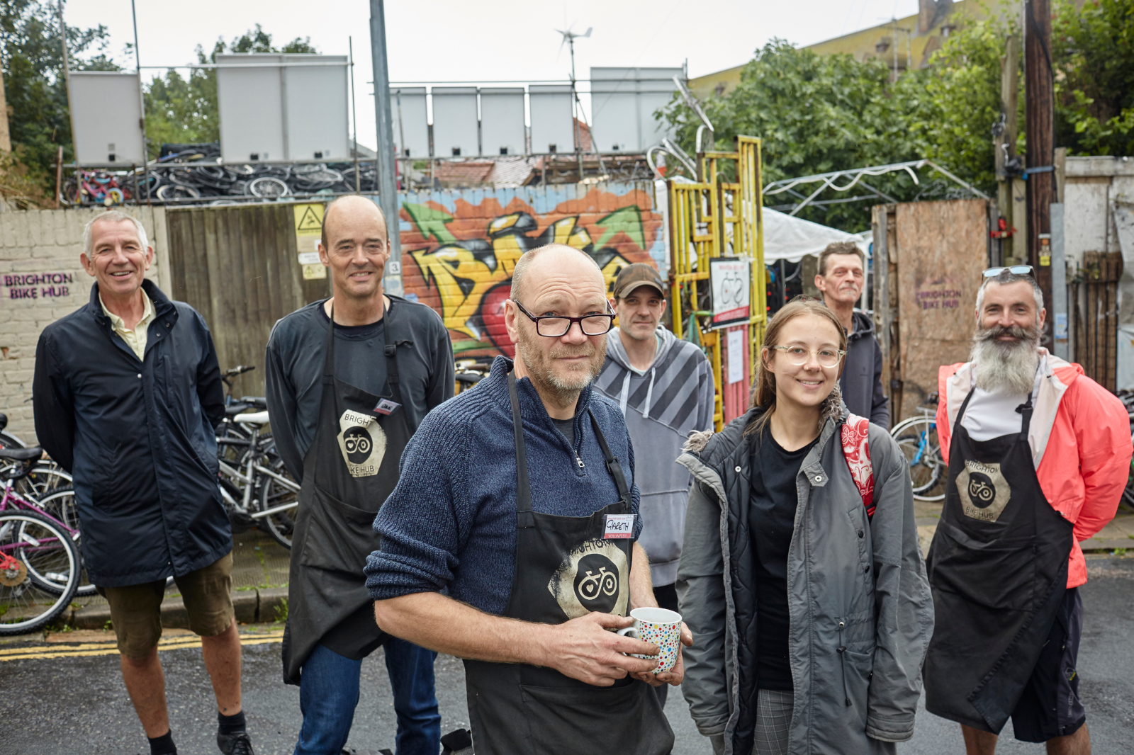 A group of individuals in aprons stands outside a bike hub, amidst bicycles and vibrant graffiti.