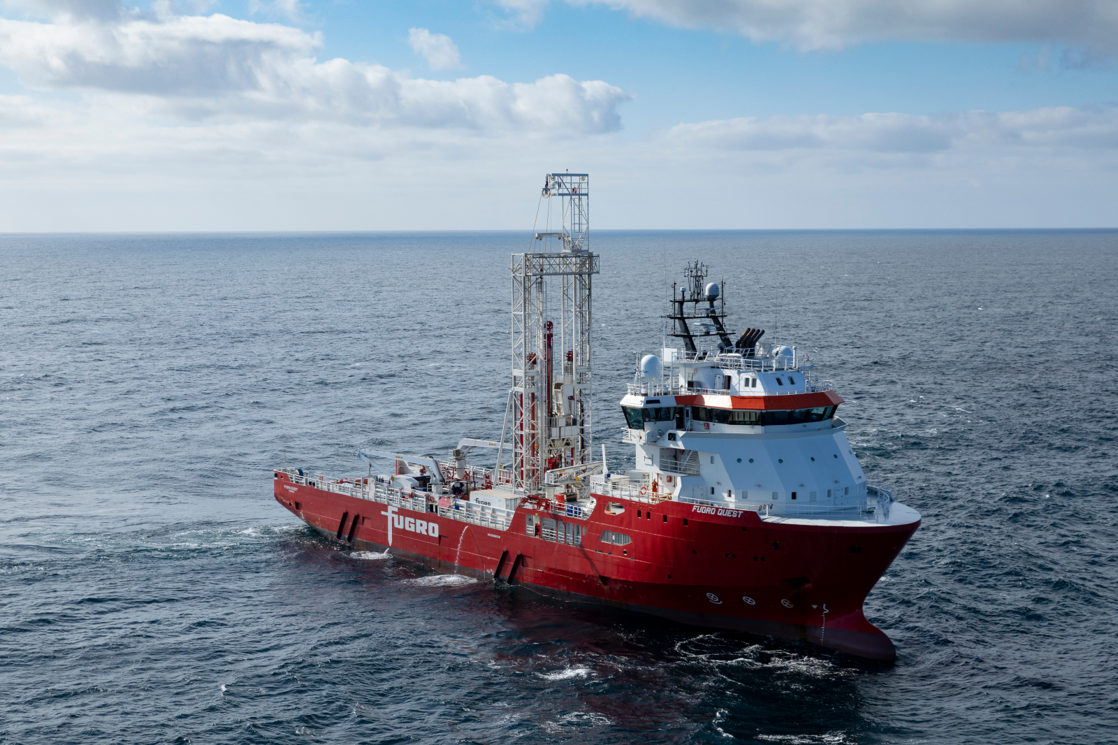 A red vessel named Fugro Quest navigates the sea, featuring drilling equipment and a clear sky above.