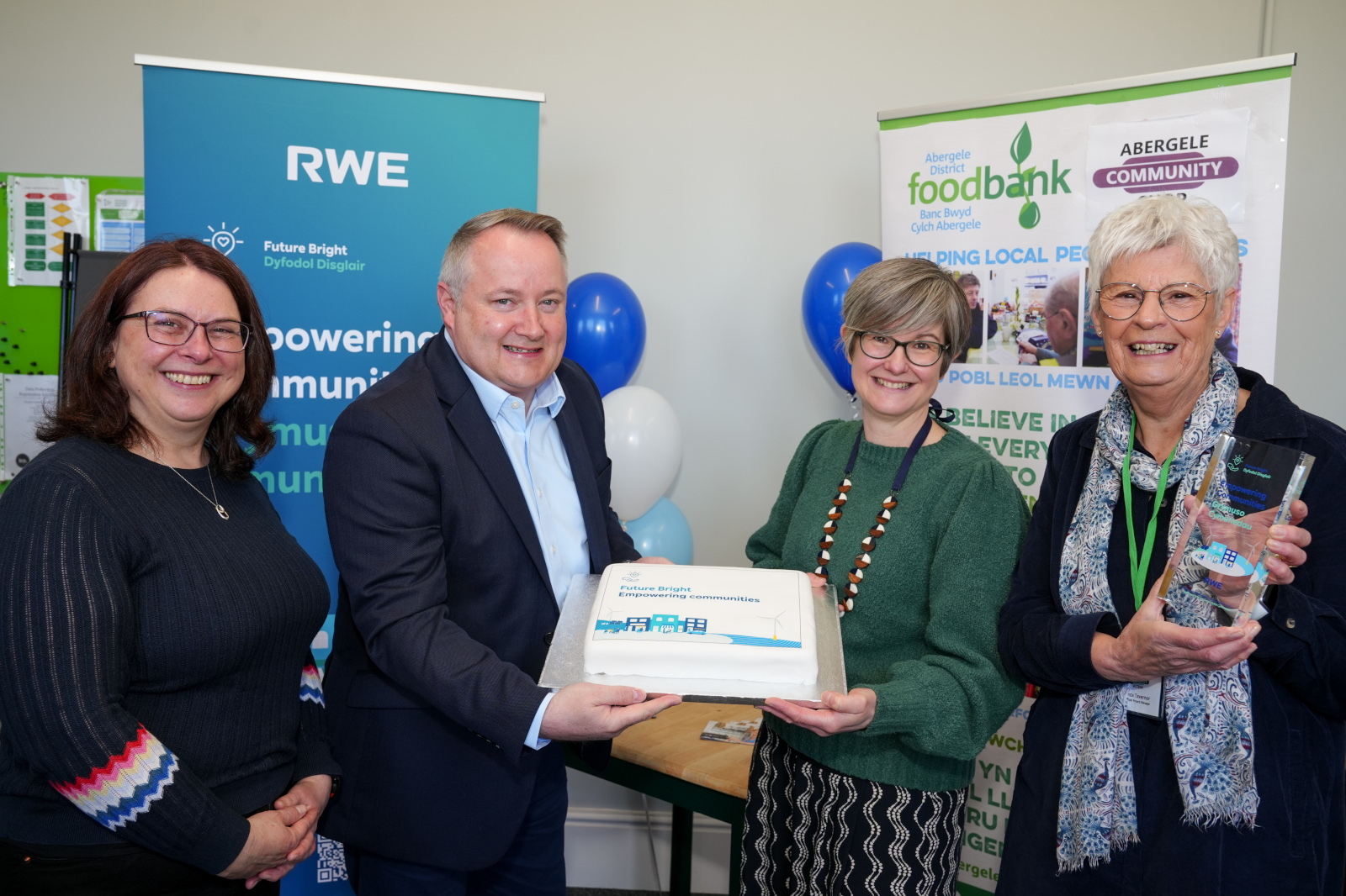 A group of individuals celebrating with a cake and awards, promoting community empowerment. Balloons and banners are visible in the background.