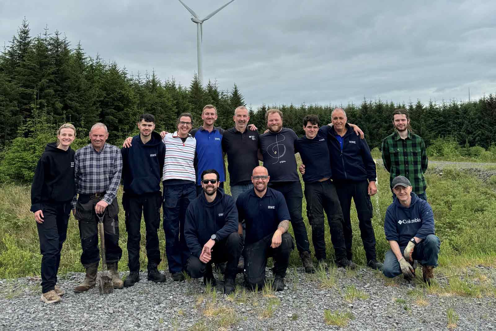 A group of eleven people stands in front of a wind turbine, surrounded by trees, with varying clothing styles and poses.