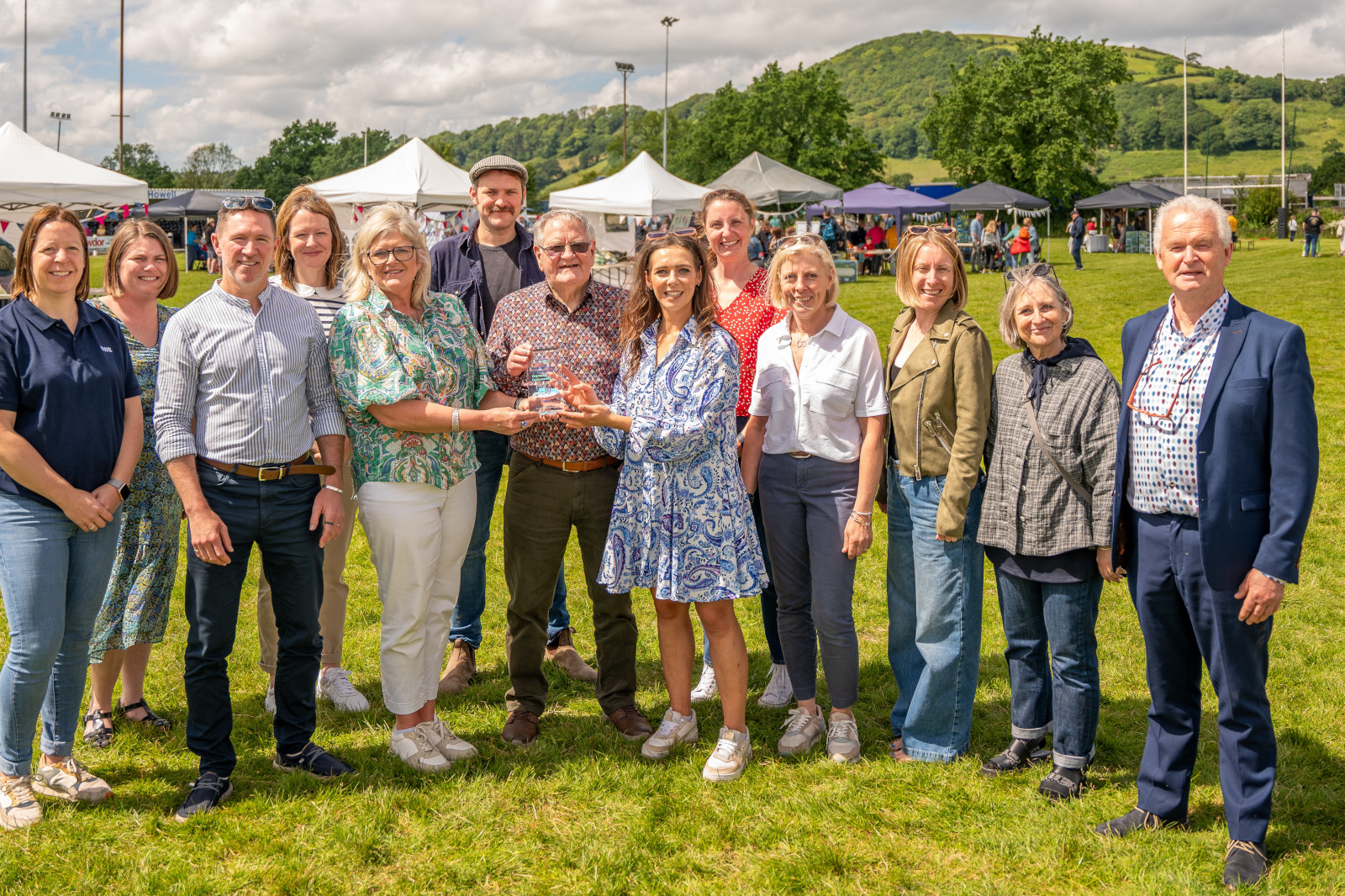 A group of people in a park holding a trophy, surrounded by tents and greenery, celebrating an event.