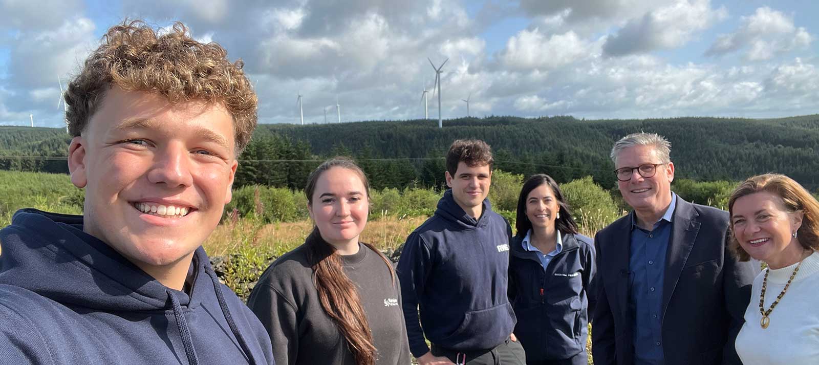 A group of six individuals stands outdoors, with lush greenery and wind turbines in the background under a partly cloudy sky.