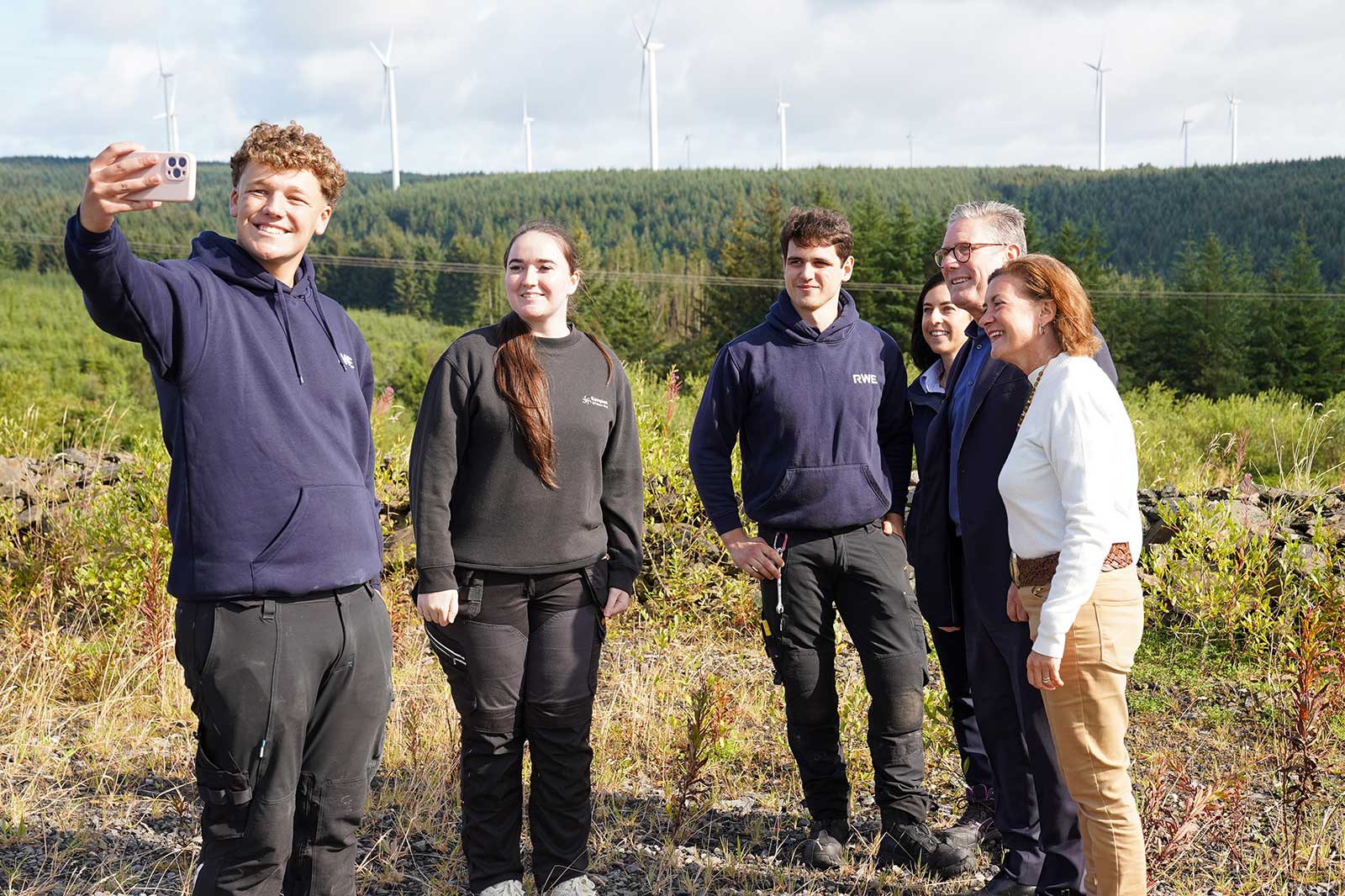 A group of people taking a selfie outdoors with a backdrop of wind turbines and lush greenery.