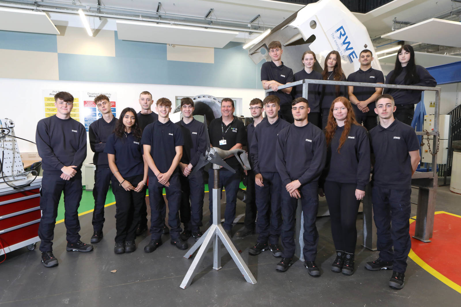 A group of individuals in black uniforms, gathered in a workshop, with machinery in the background and a model on display.