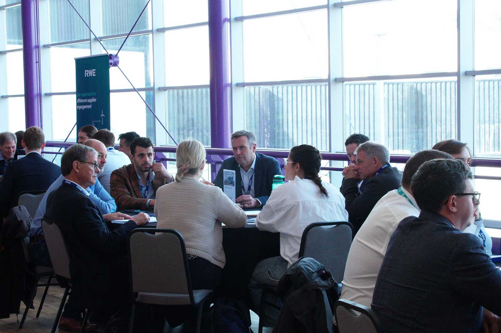 A group of professionals engaged in discussion around a round table, with a backdrop of large windows and a green branded banner.