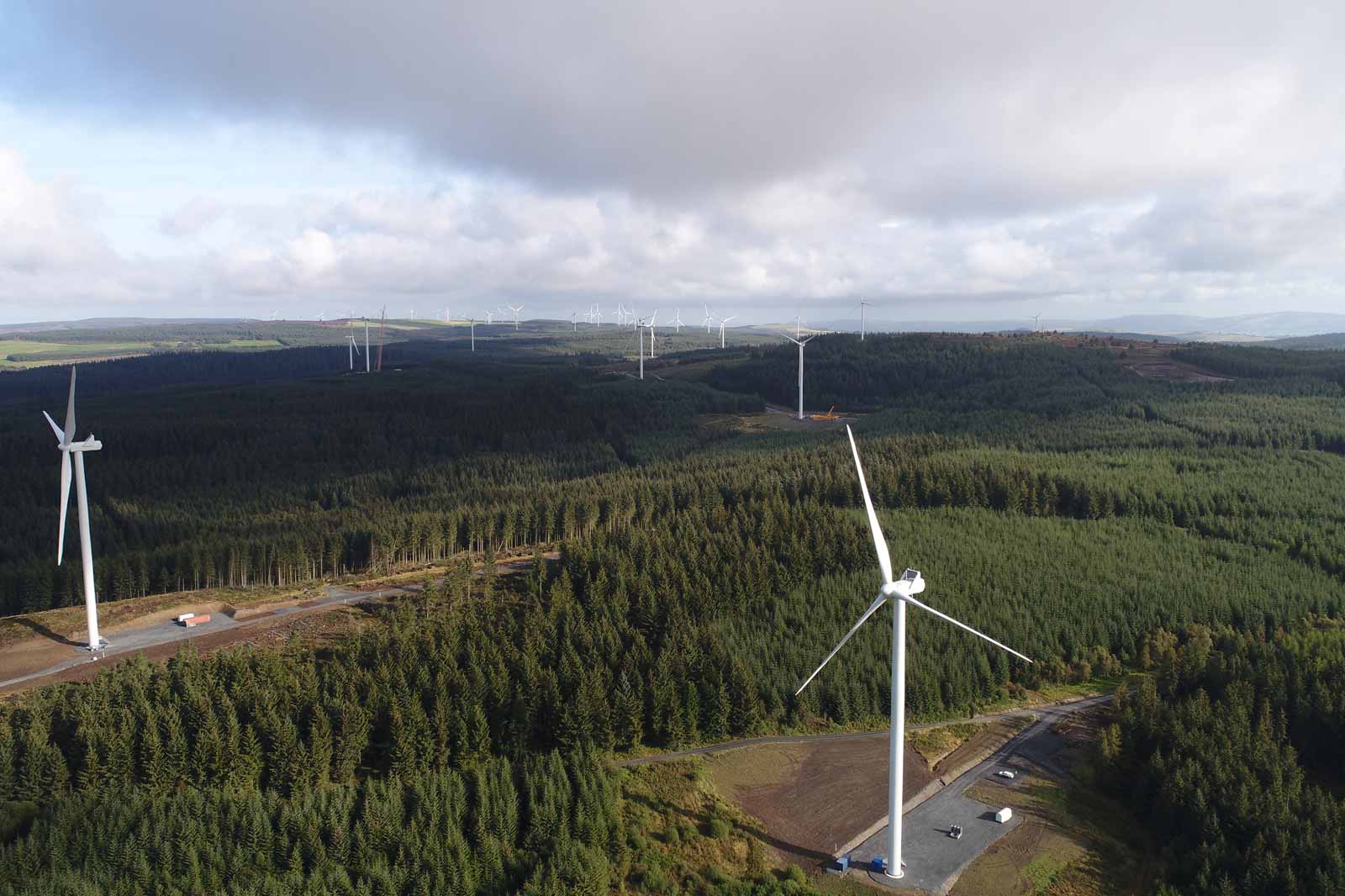 Aerial view of a wind farm with multiple turbines surrounded by dense green forests under a cloudy sky.