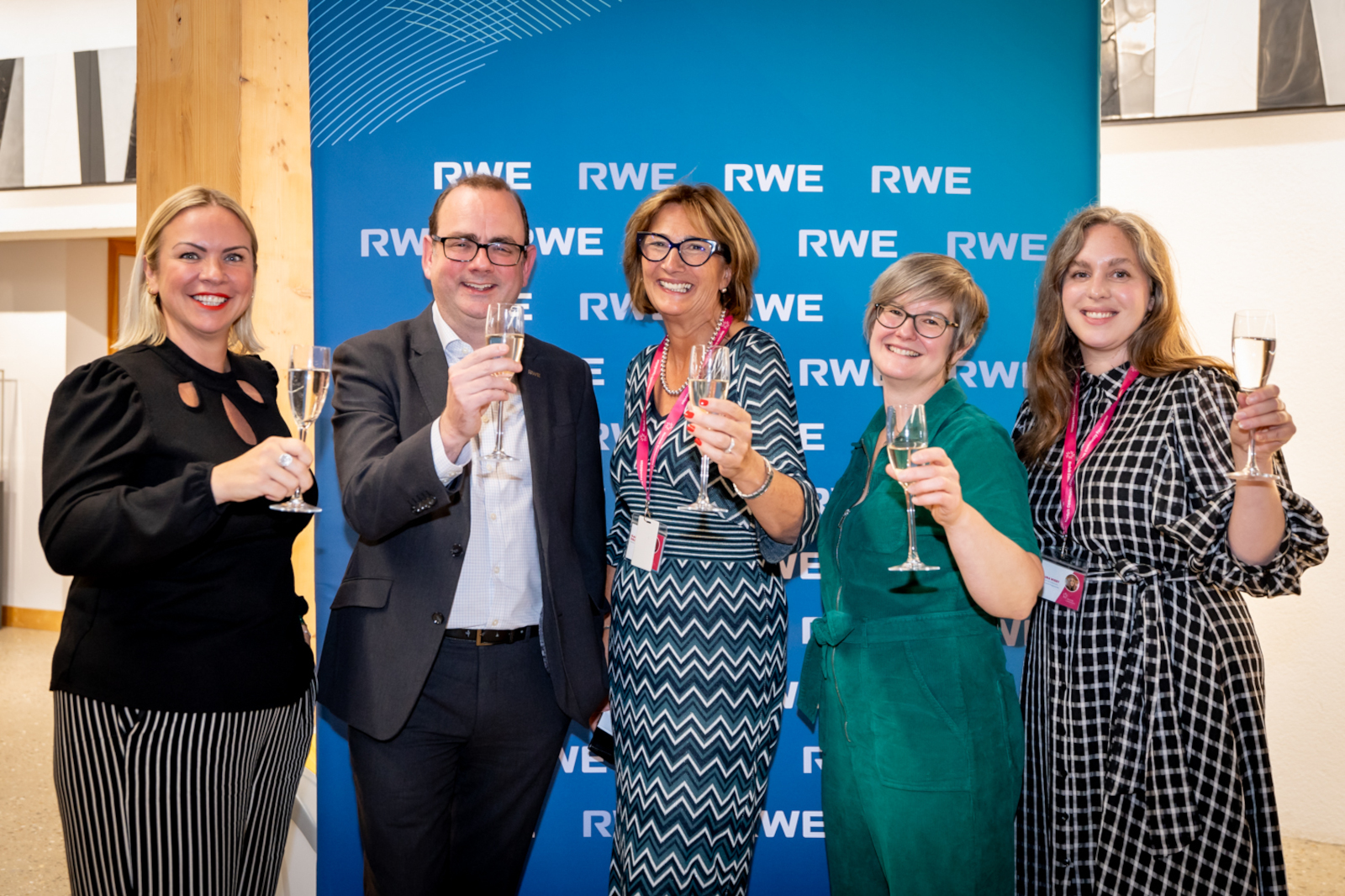 Five individuals are celebrating with glasses raised, standing in front of an RWE blue backdrop at an indoor event.