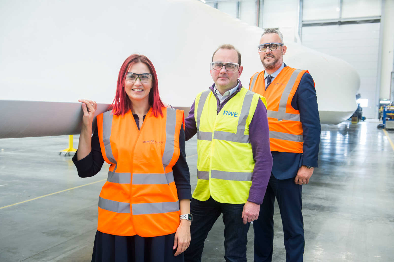 Three people in safety vests stand in a factory. A large wind turbine blade is visible in the background.