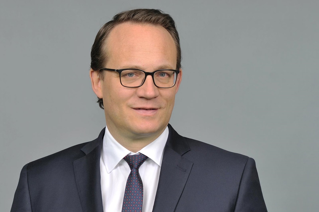 A man in a dark suit and patterned tie poses against a neutral background, conveying a professional appearance.