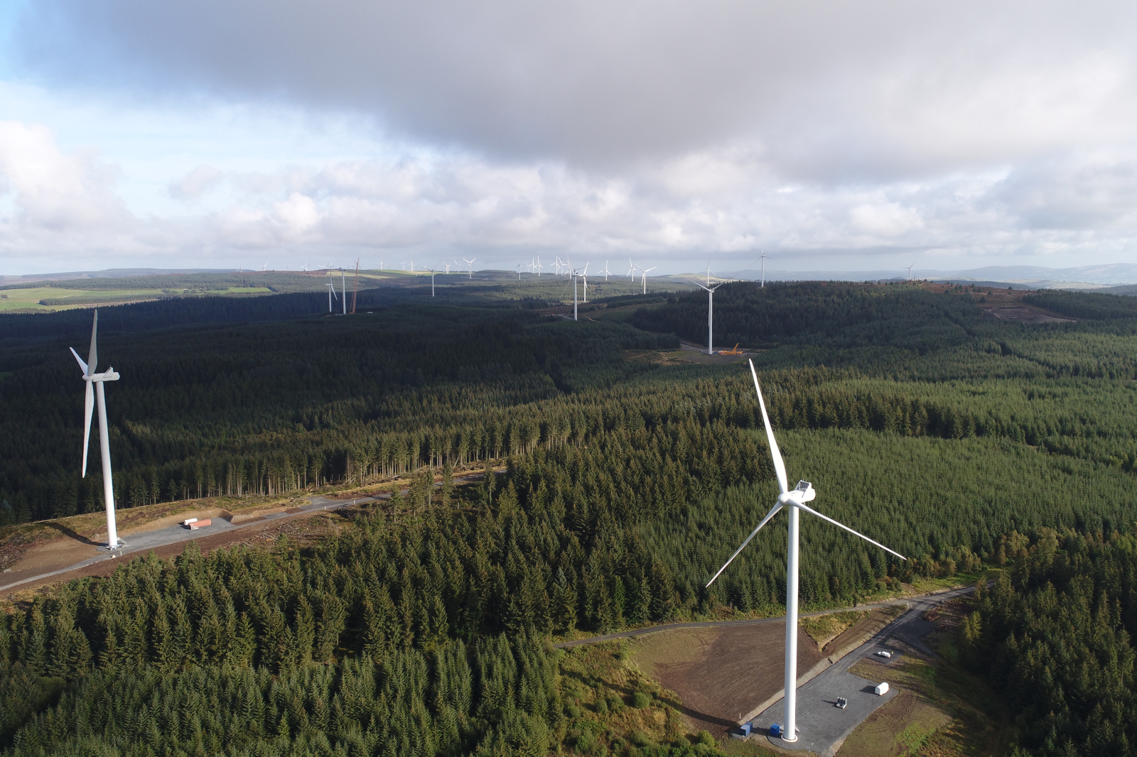 Aerial view of a wind farm with multiple turbines set against a green forest landscape under a partly cloudy sky.