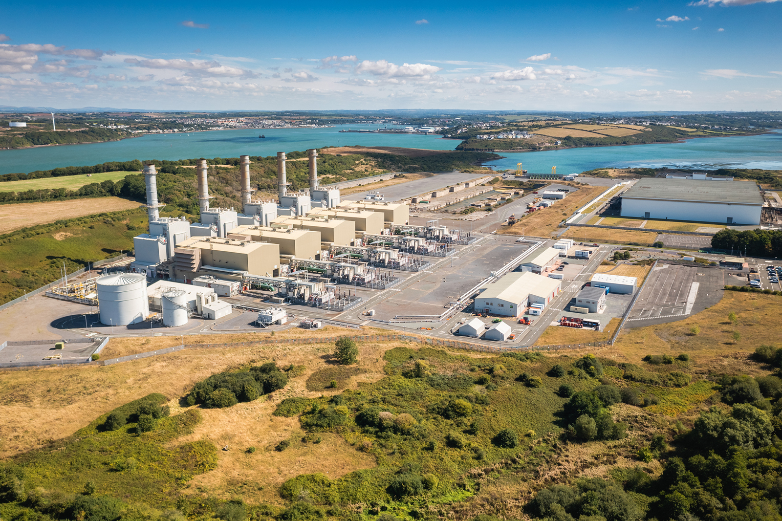 Aerial view of a power plant with smoke stacks, adjacent to a river and grassy landscape, featuring industrial buildings and storage.