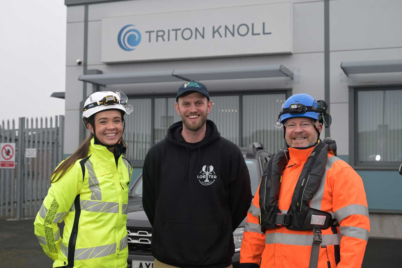 Three individuals in safety gear stand outside the Triton Knoll facility, with a grey vehicle and a fence in the background.