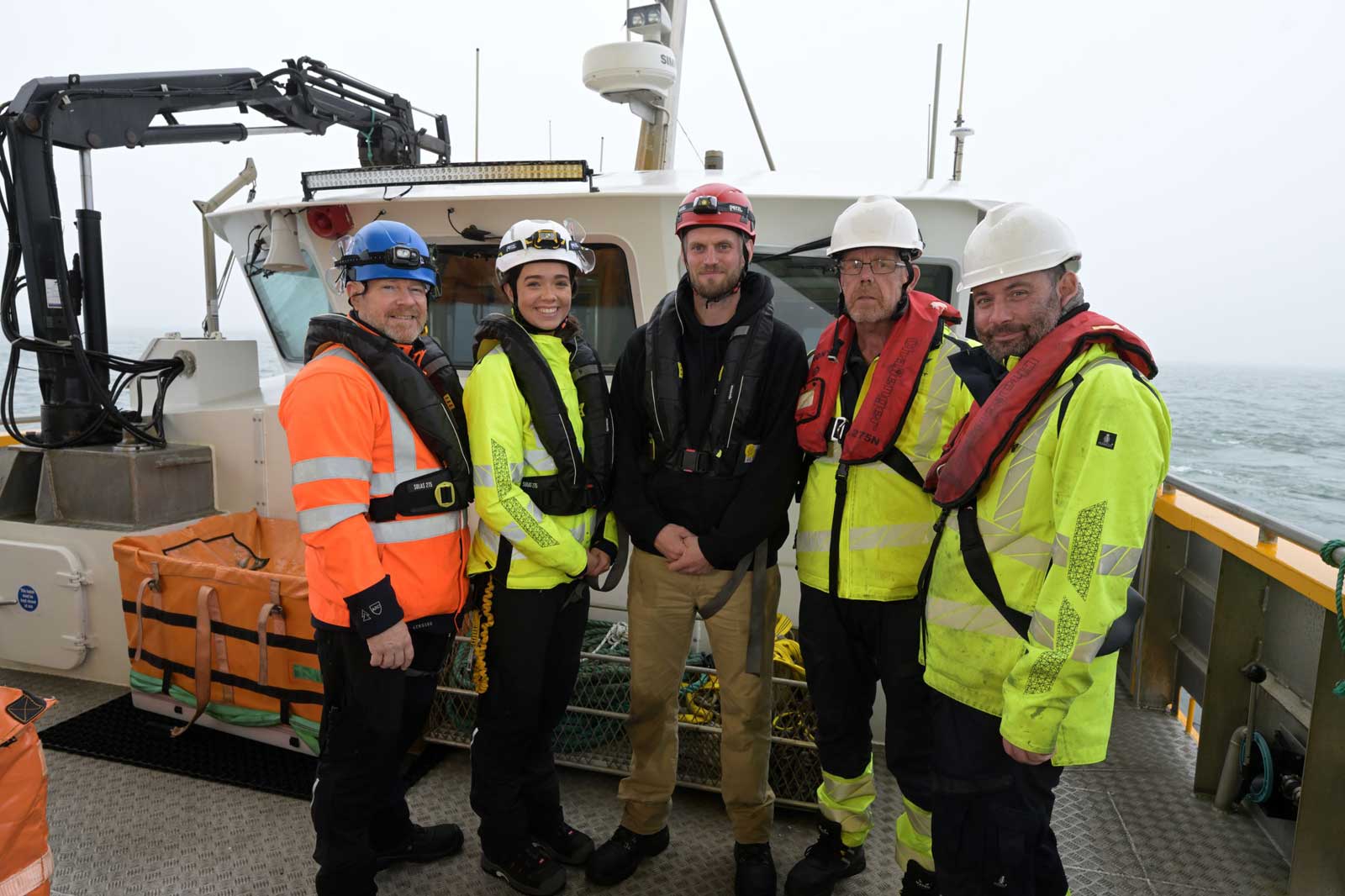 Five crew members in safety gear stand on a boat, with a grey ocean and fog in the background.