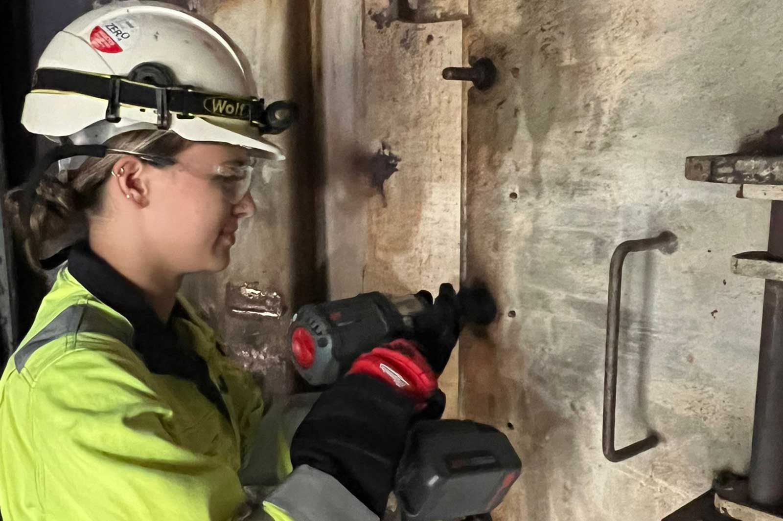 A worker in a yellow safety jacket uses a power drill on a metal surface in an industrial setting.