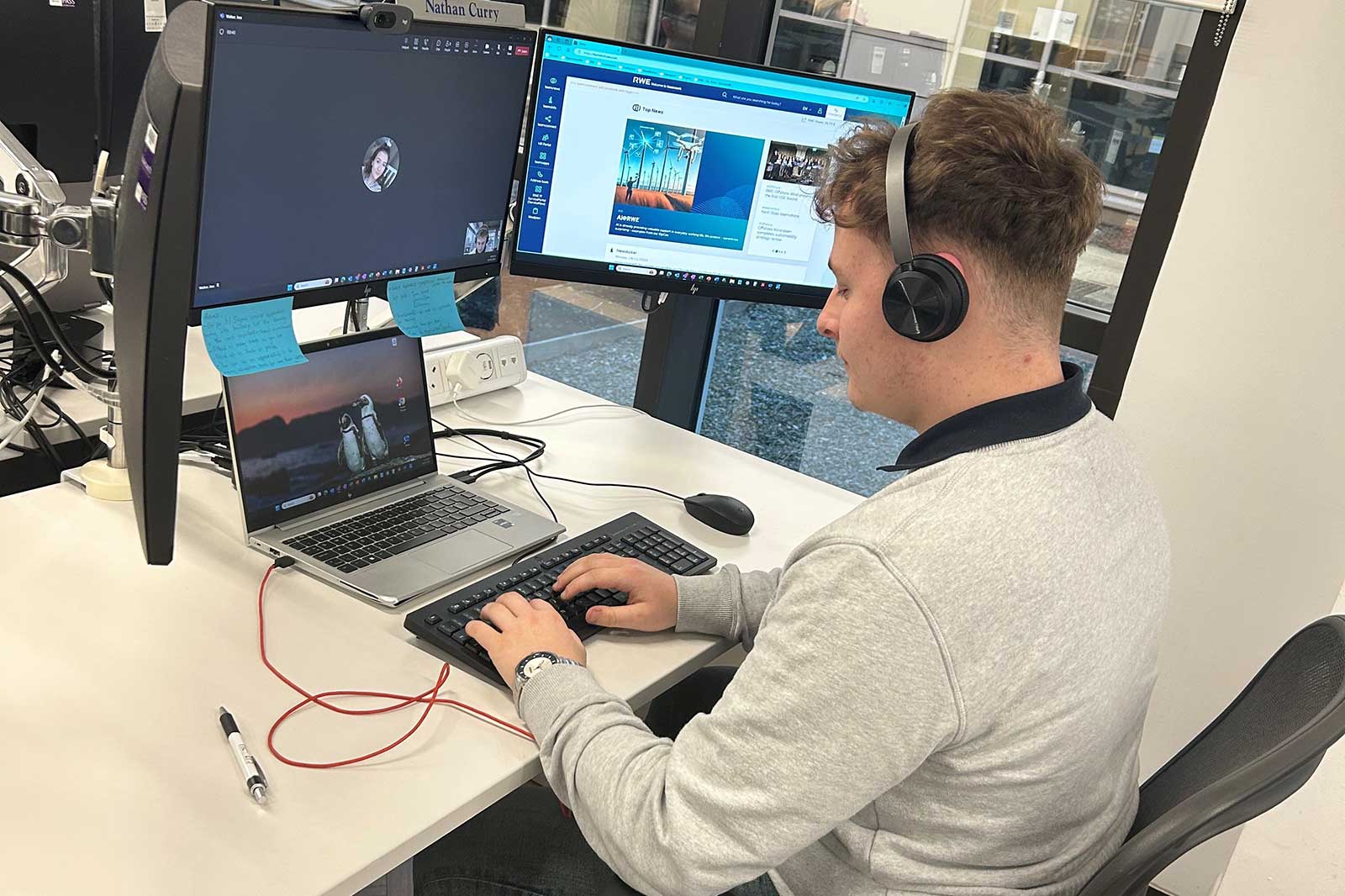 A person working at a desk with two computer monitors, headphones, and a laptop displaying a penguin image.