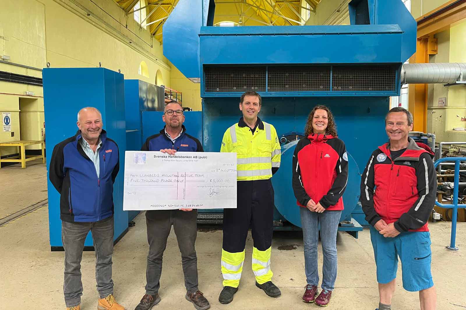 Group of four individuals holding a large cheque in front of industrial machinery, showcasing support for a rescue team.