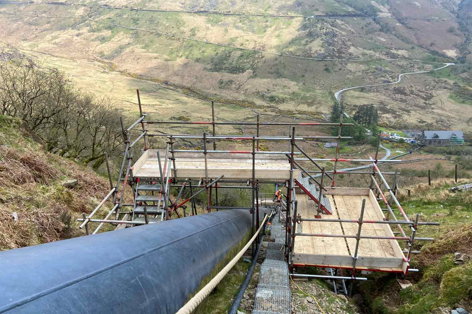 A construction site showing scaffolding around a large pipe on a hillside, with a worker in an orange safety vest.