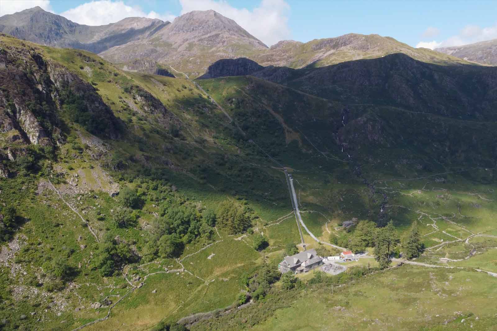 A scenic view of a valley surrounded by mountains, featuring a small building with a red roof and lush greenery.