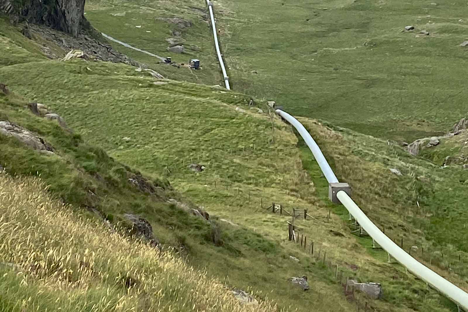 A long white pipe runs through a grassy hillside, surrounded by rocks and sparse vegetation under a cloudy sky.