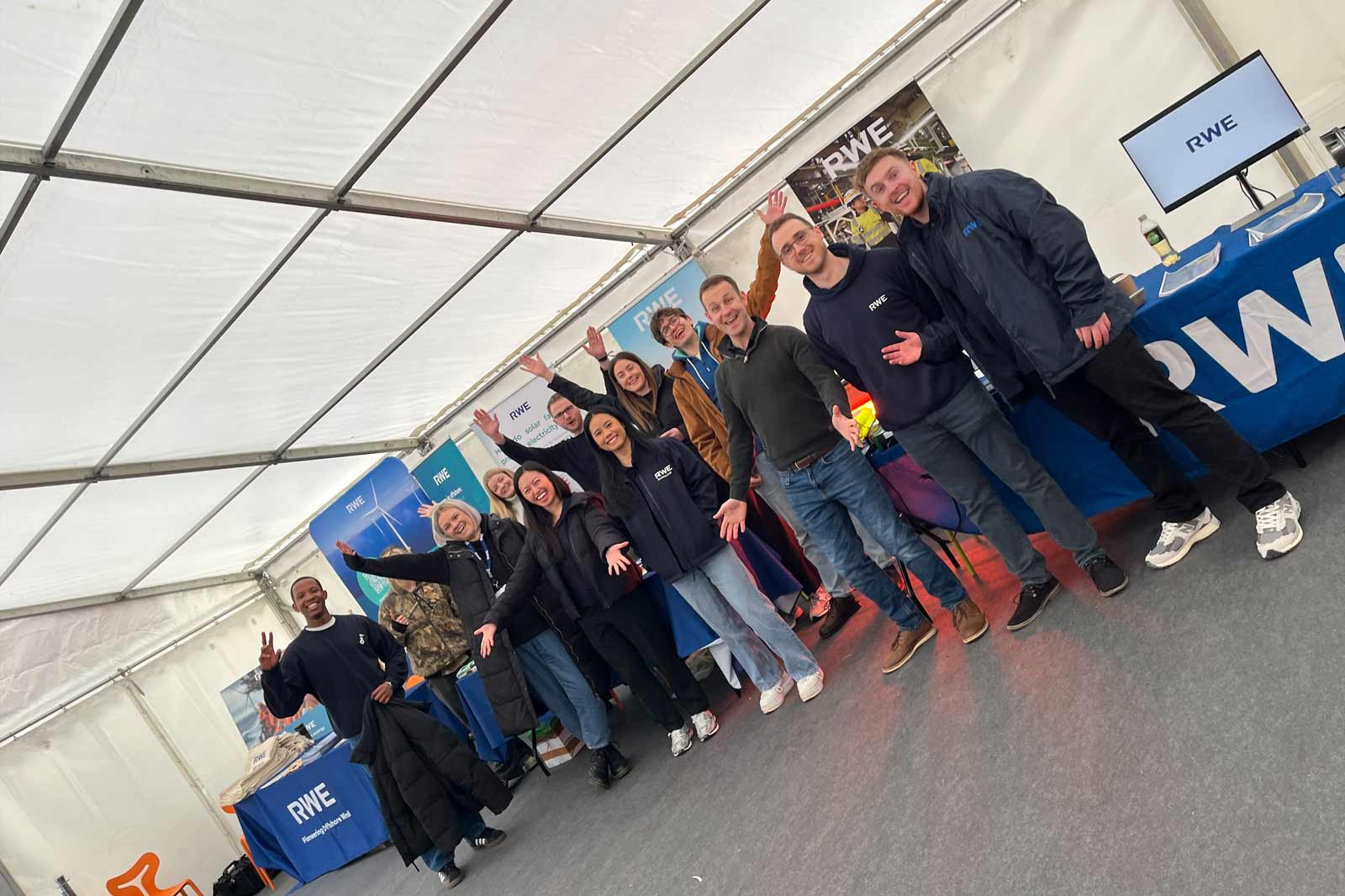 A group of people cheerfully posing indoors, wearing RWE branded jackets, with promotional materials and a screen visible in the background.