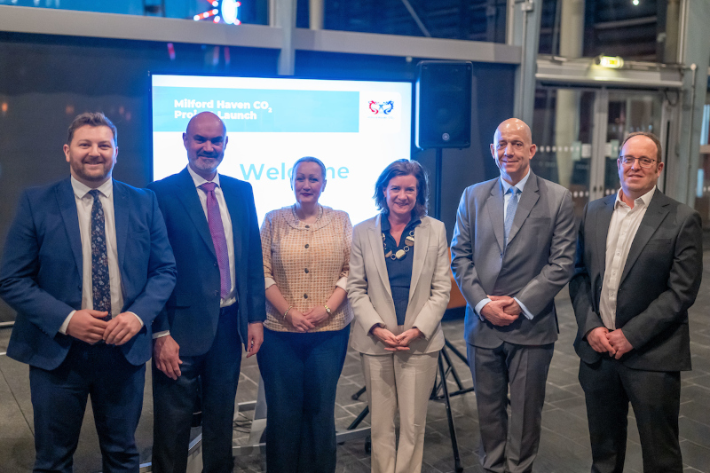 A group of six professionals stands together at the Milford Haven CO2 project launch, smiling in front of a welcome sign.