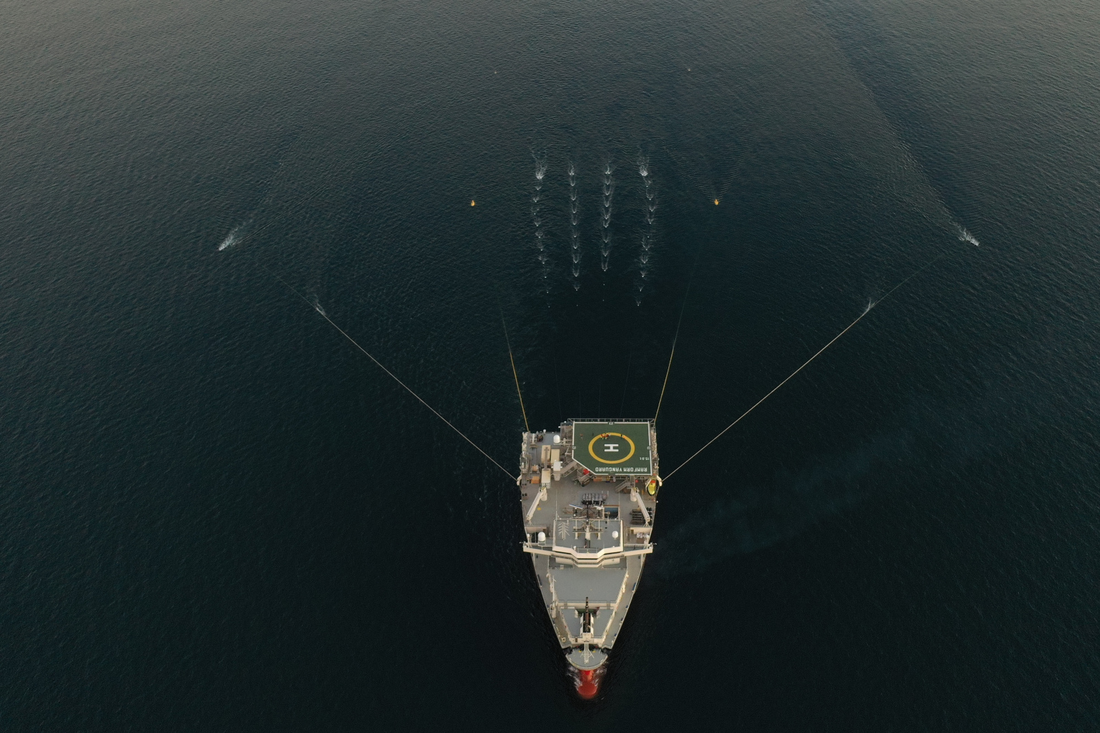 An aerial view of a ship with a helipad in the ocean, trailing lines and producing spray on the water surface.