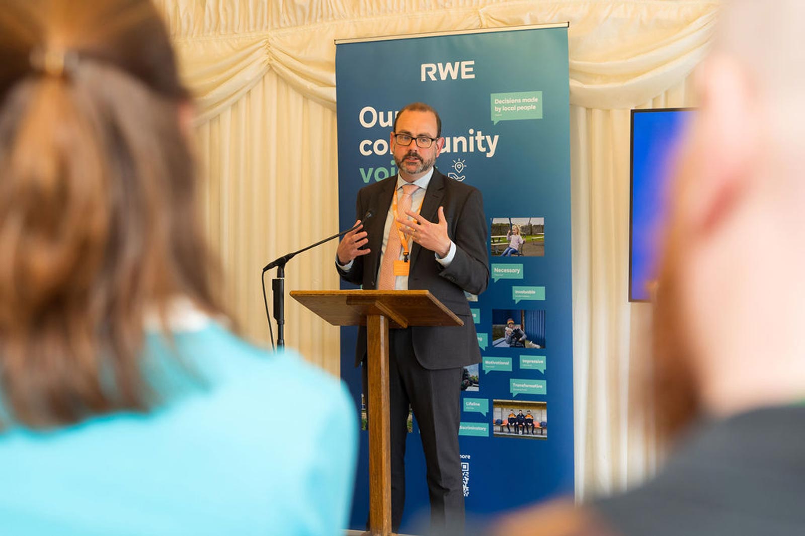 A speaker presents at an event with an RWE banner in the background, engaging the audience while standing at a wooden podium.