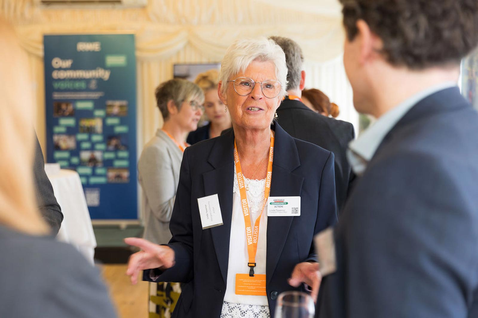 A woman in a dark blazer and white blouse gestures while talking at a networking event, surrounded by other attendees.