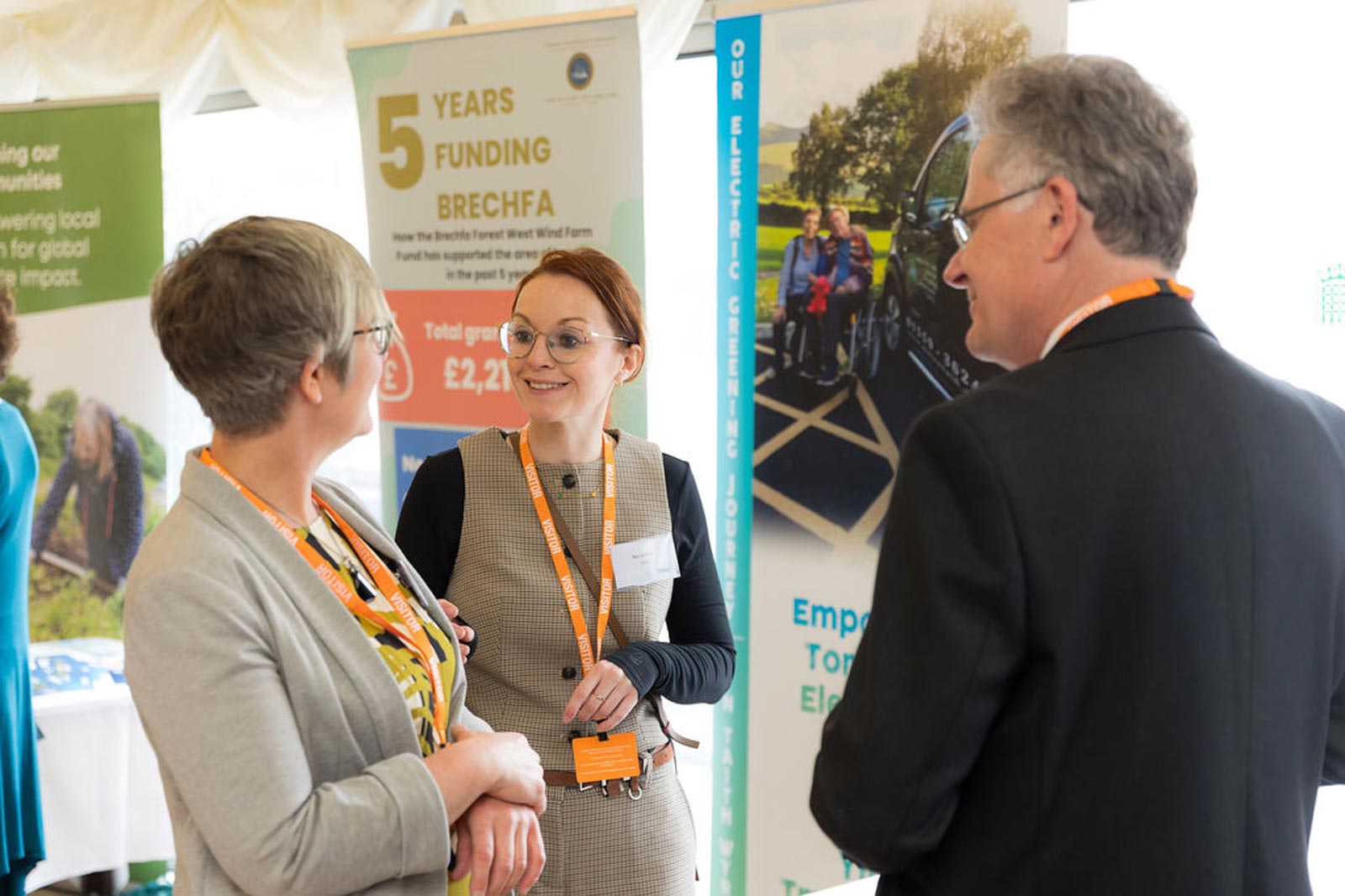 Three individuals engage in conversation near informative banners about funding and electric transportation initiatives.