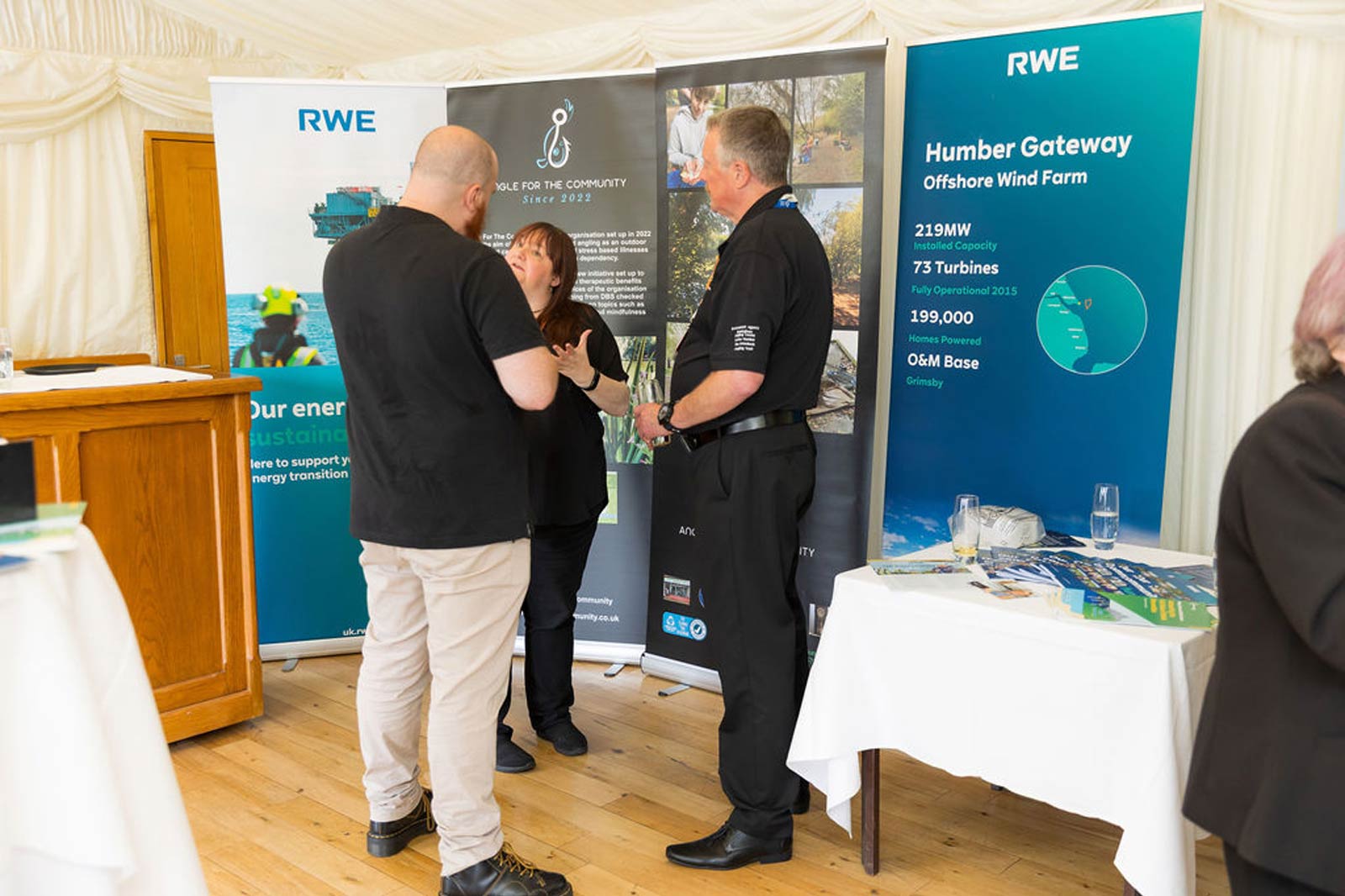Two people converse with a representative at a wind farm event. Banners display information about RWE and Humber Gateway.
