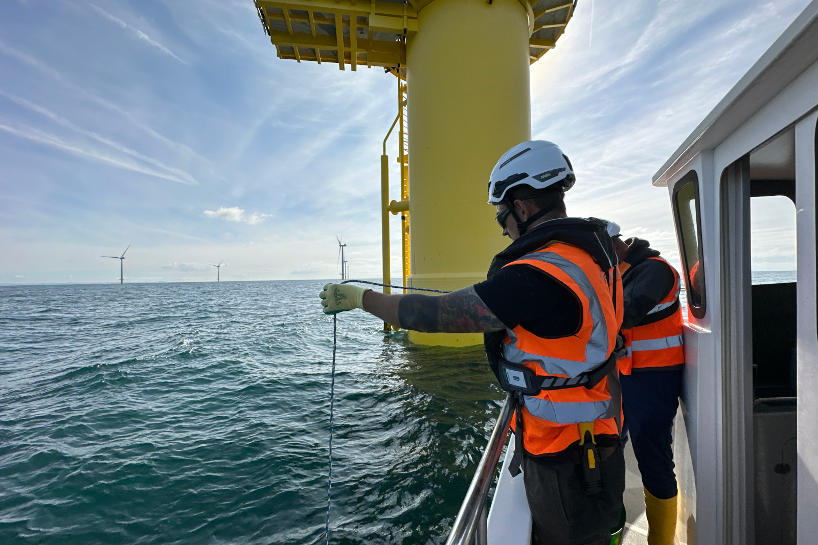 Two workers in safety vests are observing the ocean from a boat near a large yellow wind turbine structure.
