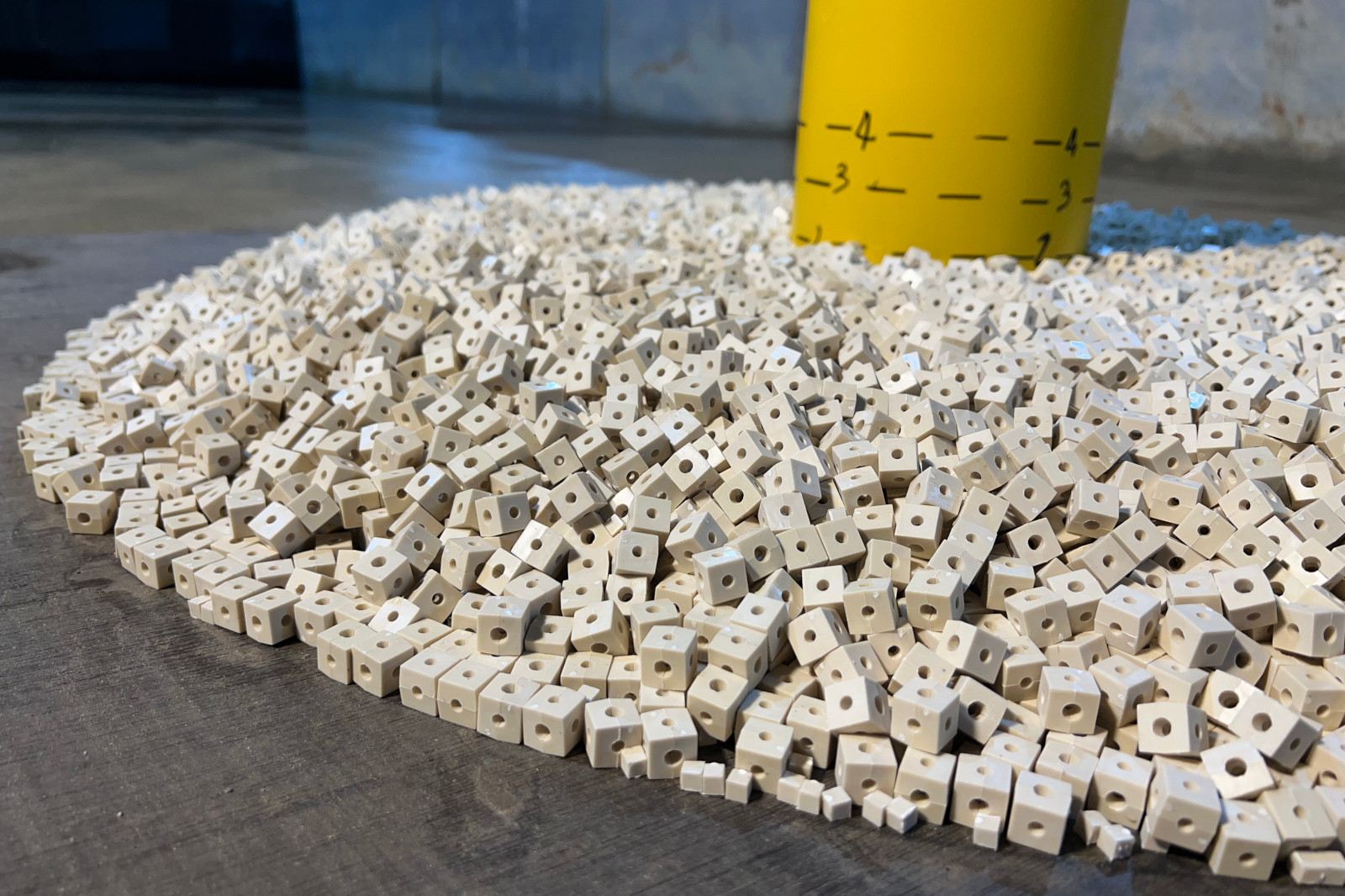 A pile of small white cubes with holes on a concrete floor, beside a yellow measuring cylinder marked with levels.