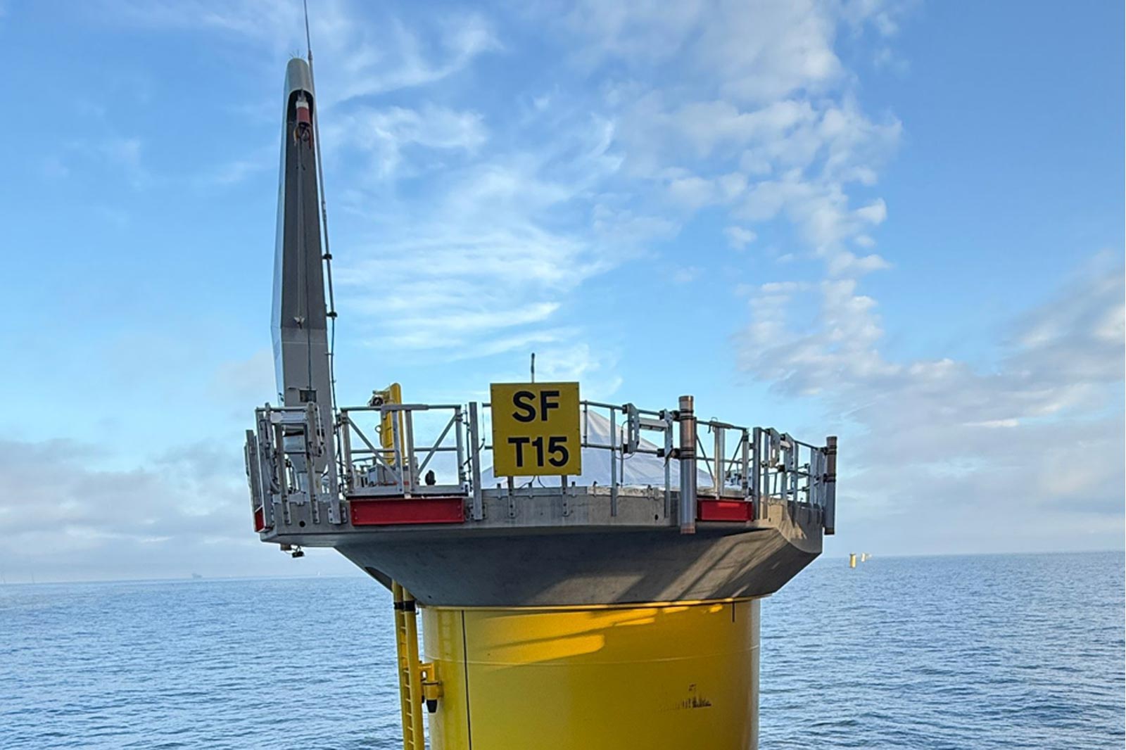 A yellow offshore wind turbine base with a platform marked SF T15 under a blue sky and calm ocean.