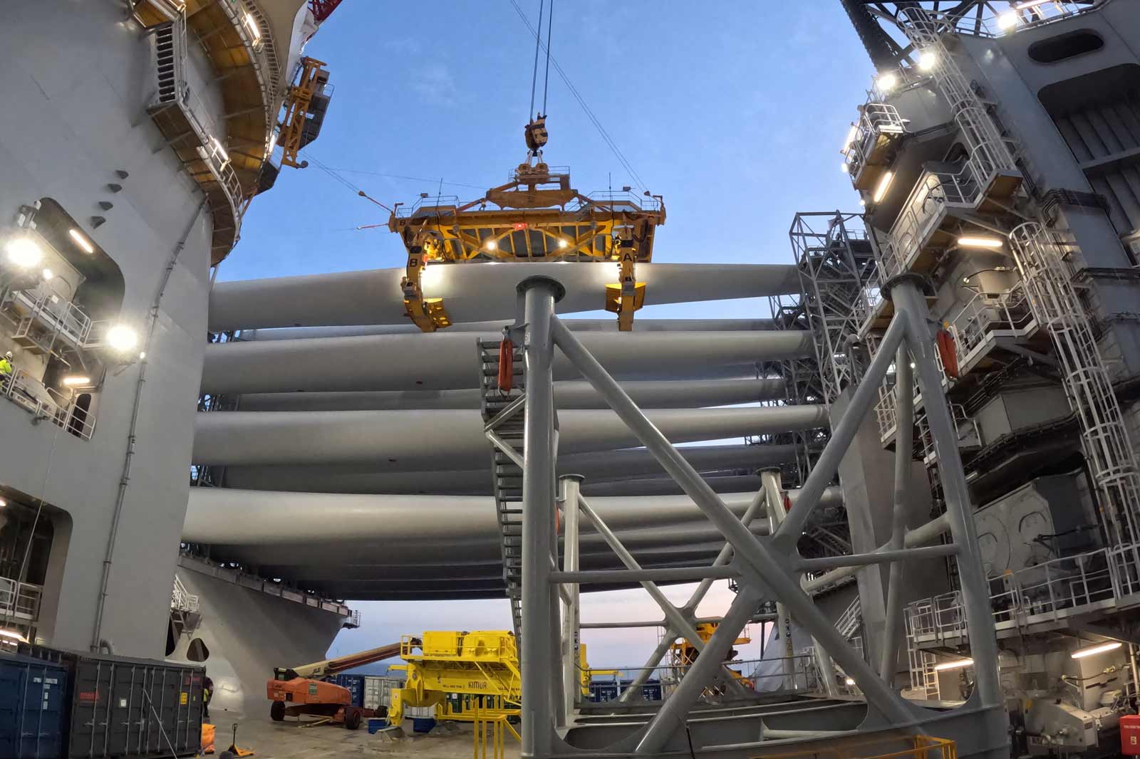 A crane lifts large pipes on an offshore platform, surrounded by various machinery and lighting.