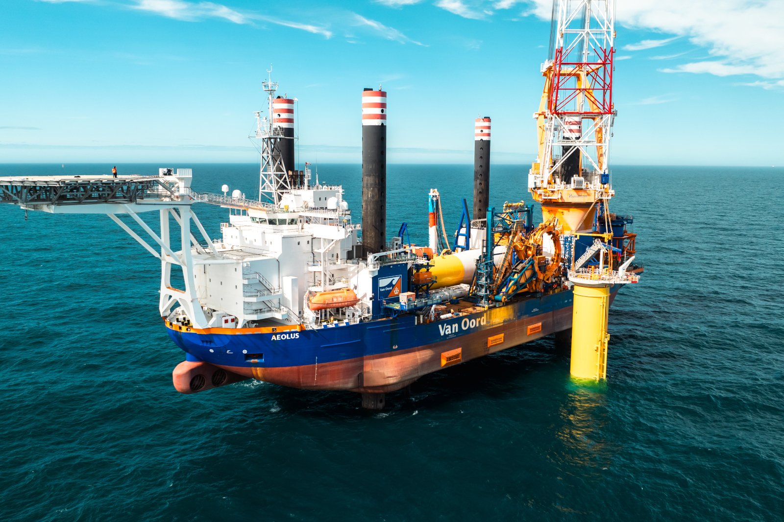 A blue and orange ship named Aeolus with four tall black smokestacks and a yellow structure, anchored in the calm sea.