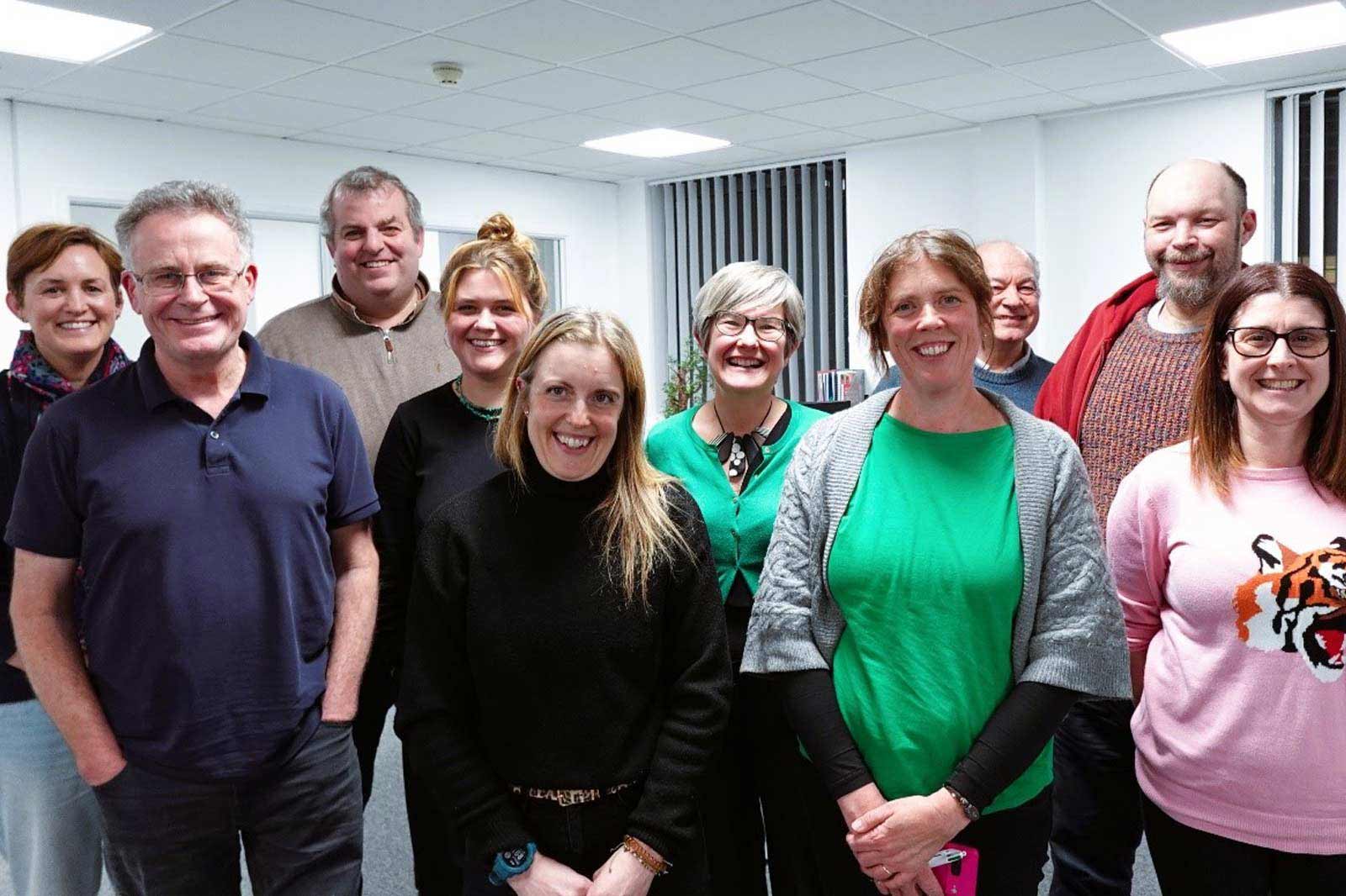 A group of individuals stands together in an indoor setting. They are wearing various outfits, predominantly in shades of green and pink.