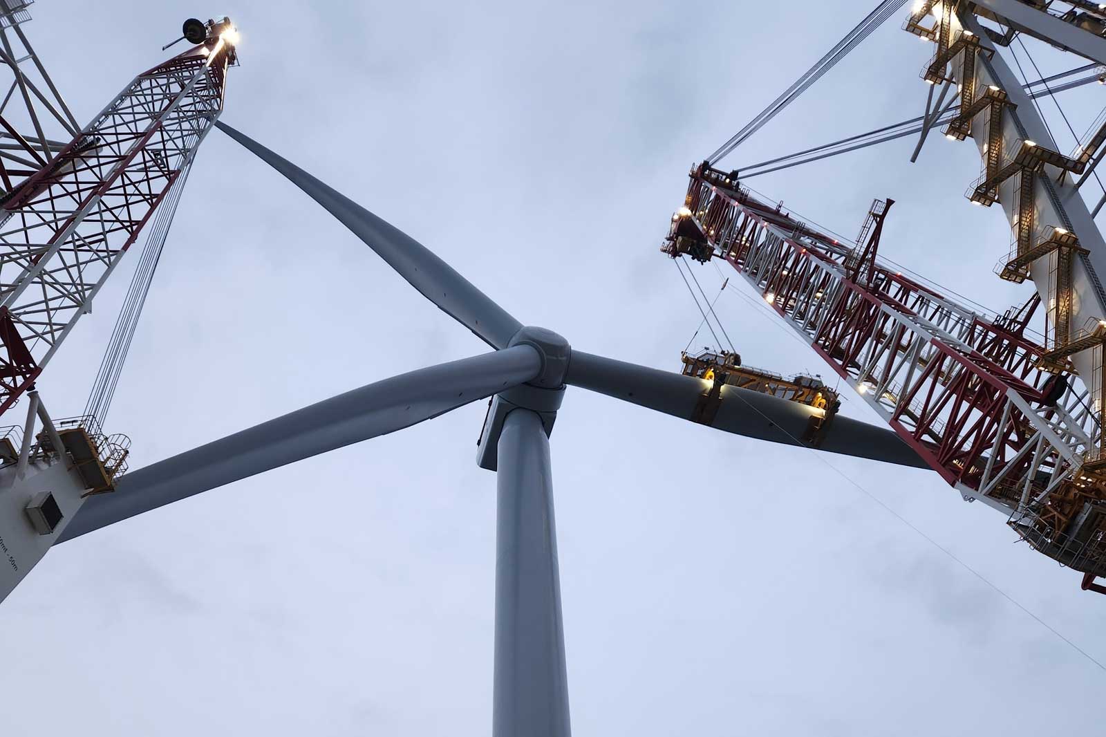 A low-angle view of a wind turbine and nearby construction cranes against a cloudy sky.