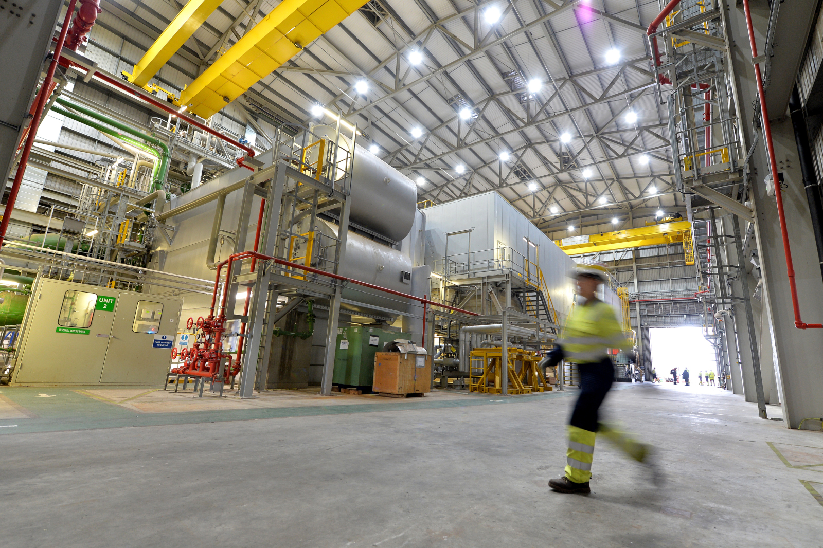 An industrial facility with large machinery, pipes, and workers in high visibility clothing walking through a spacious area.