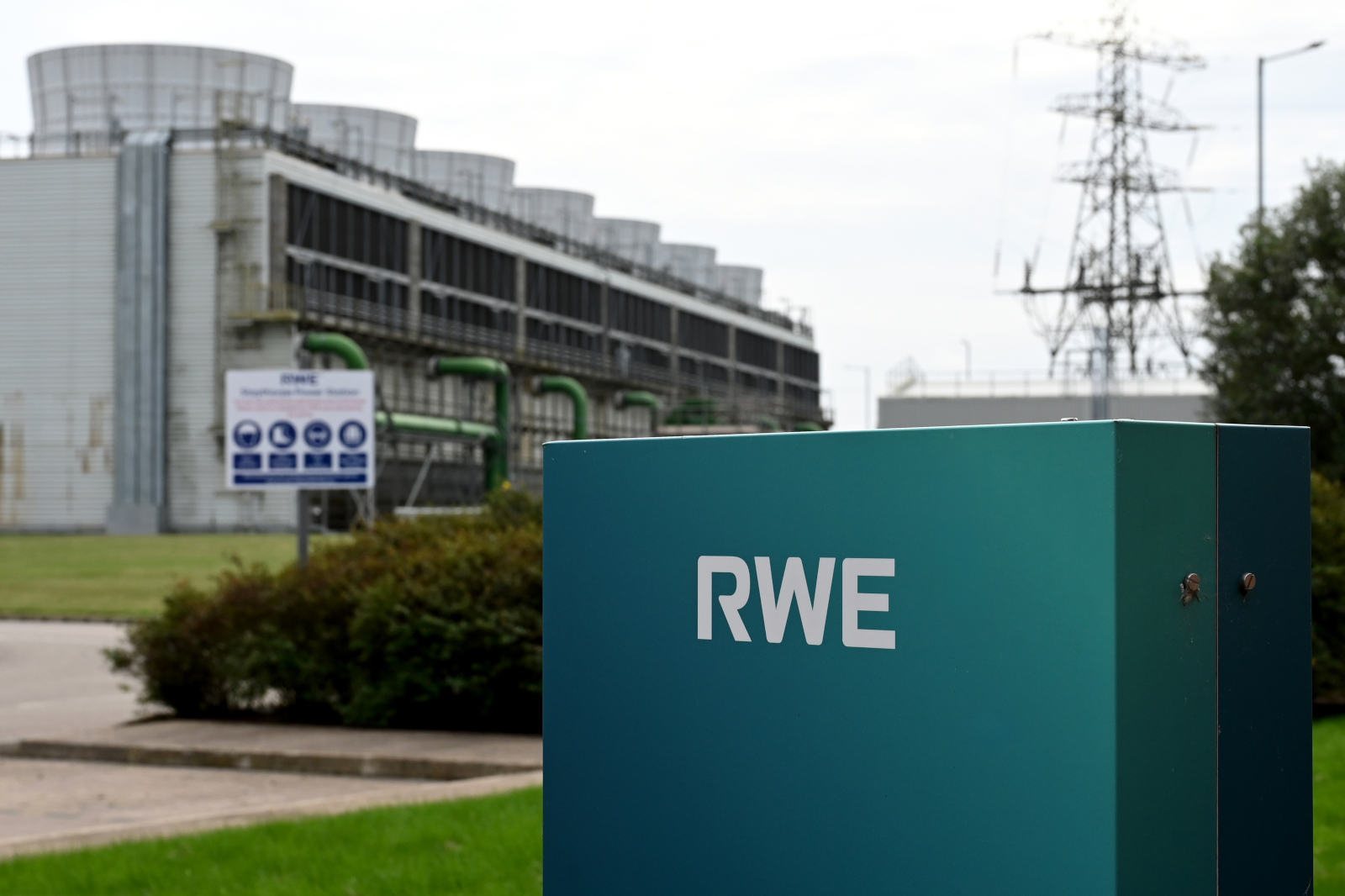 A large industrial building with cooling towers, RWE logo prominently displayed on a sign in the foreground, and green pipes.