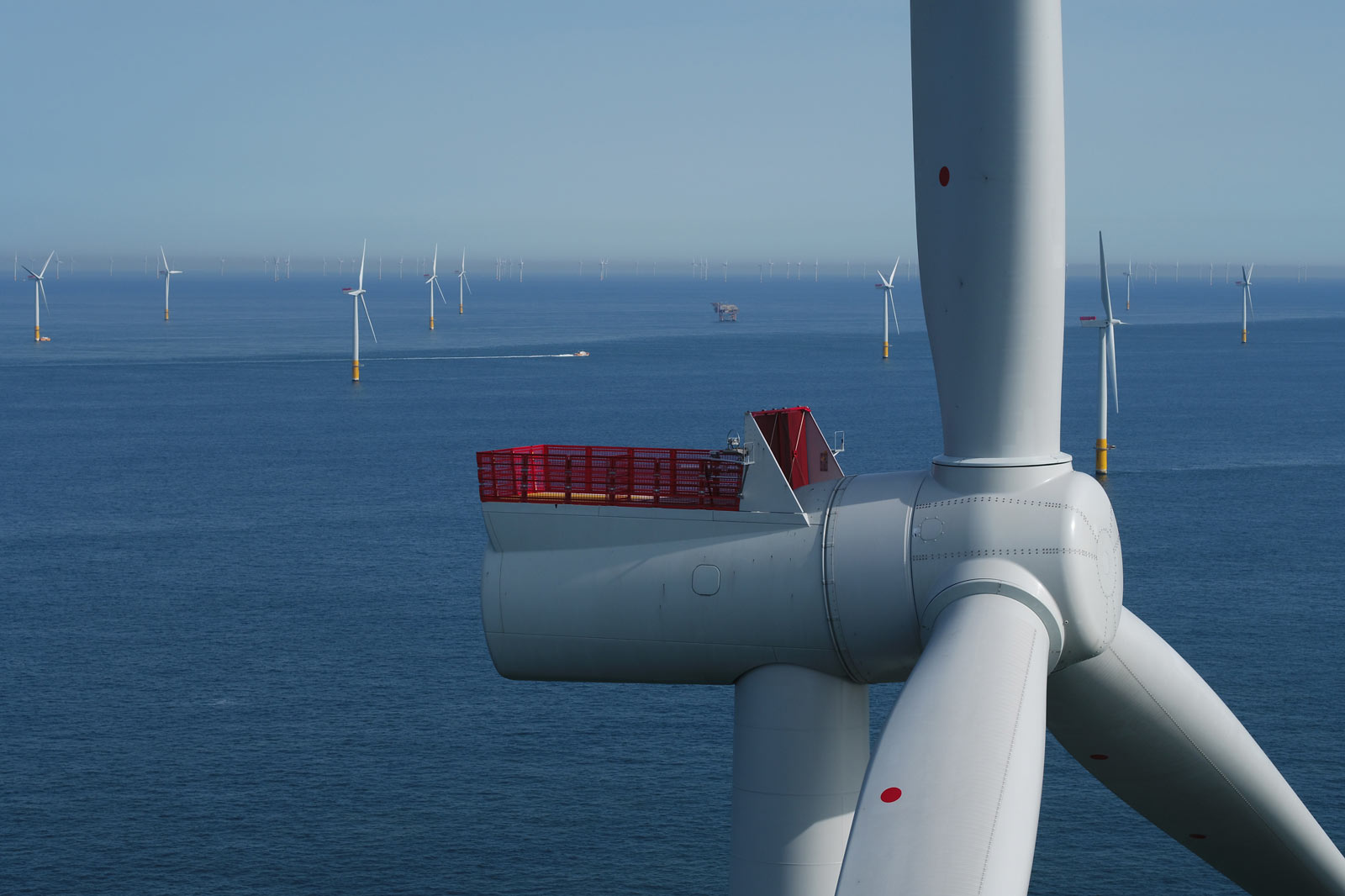 A close-up of a wind turbine with a red observation deck overlooking multiple wind turbines on the ocean in clear weather.